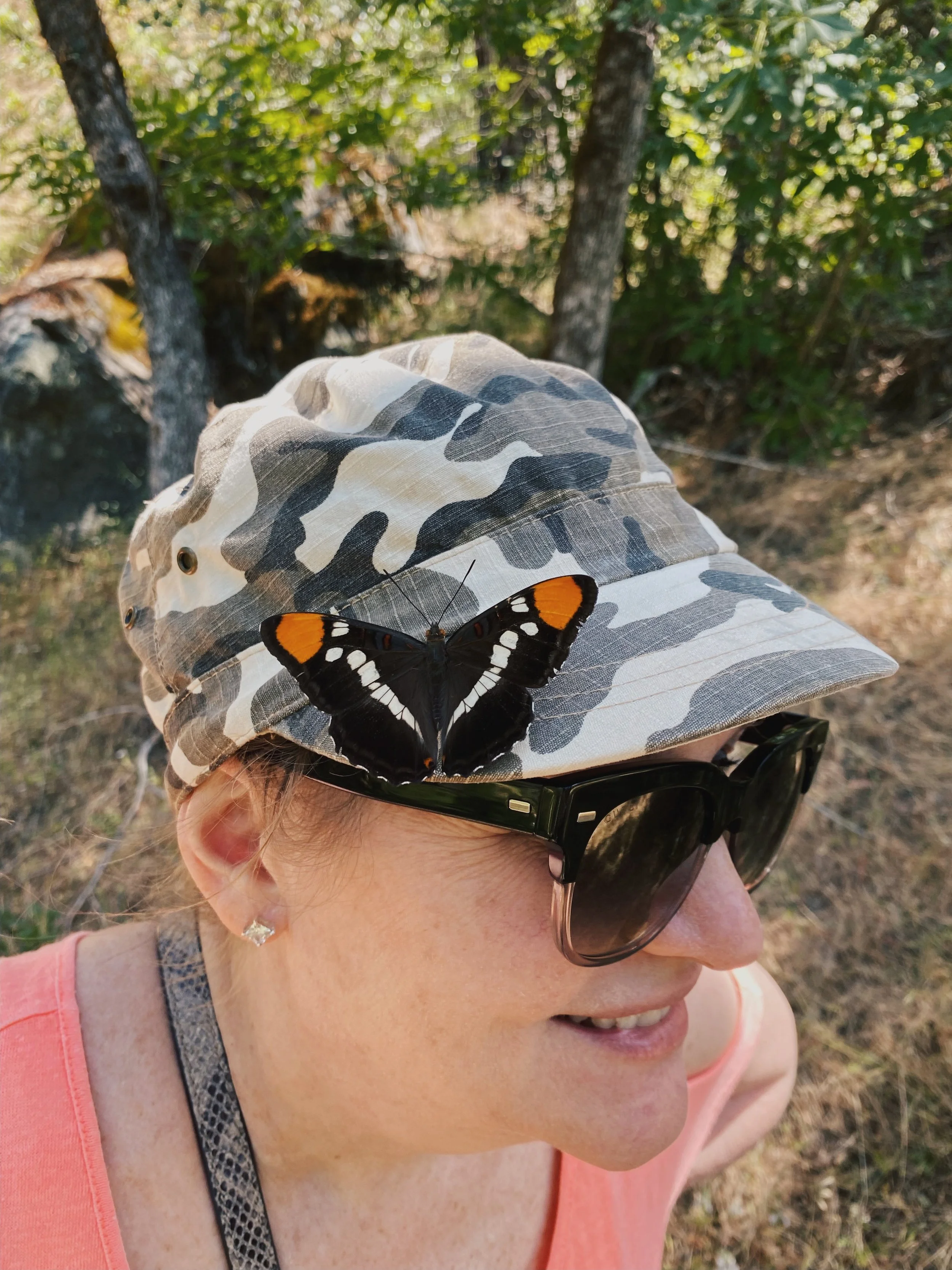 A woman wearing sunglasses and a camouflage baseball cap with a monarch butterfly resting on the brim, outdoors in a wooded area with sunlight filtering through trees.
