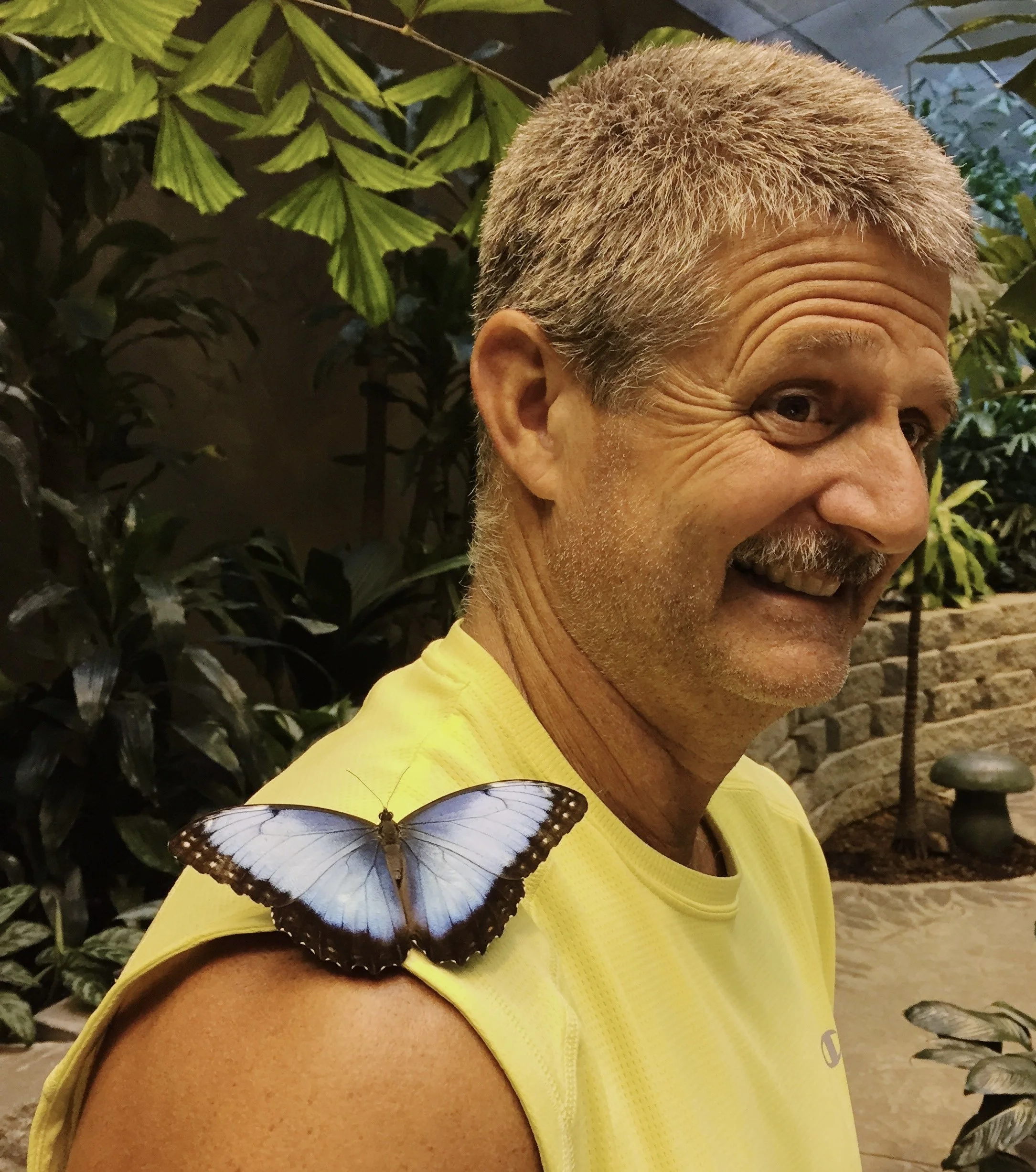 The Bug Guy in a yellow shirt with a butterfly perched on his shoulder, smiling and looking to the side amidst green plants.