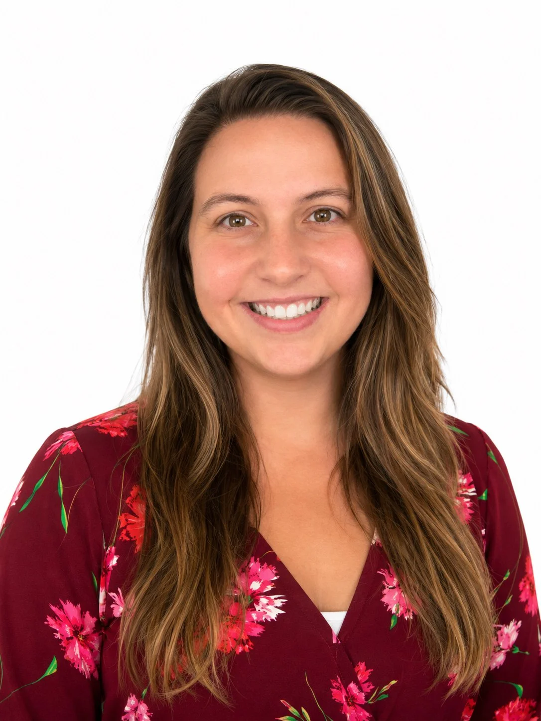 Portrait of a young woman with long brown hair, smiling, wearing a red floral blouse, against a white background.