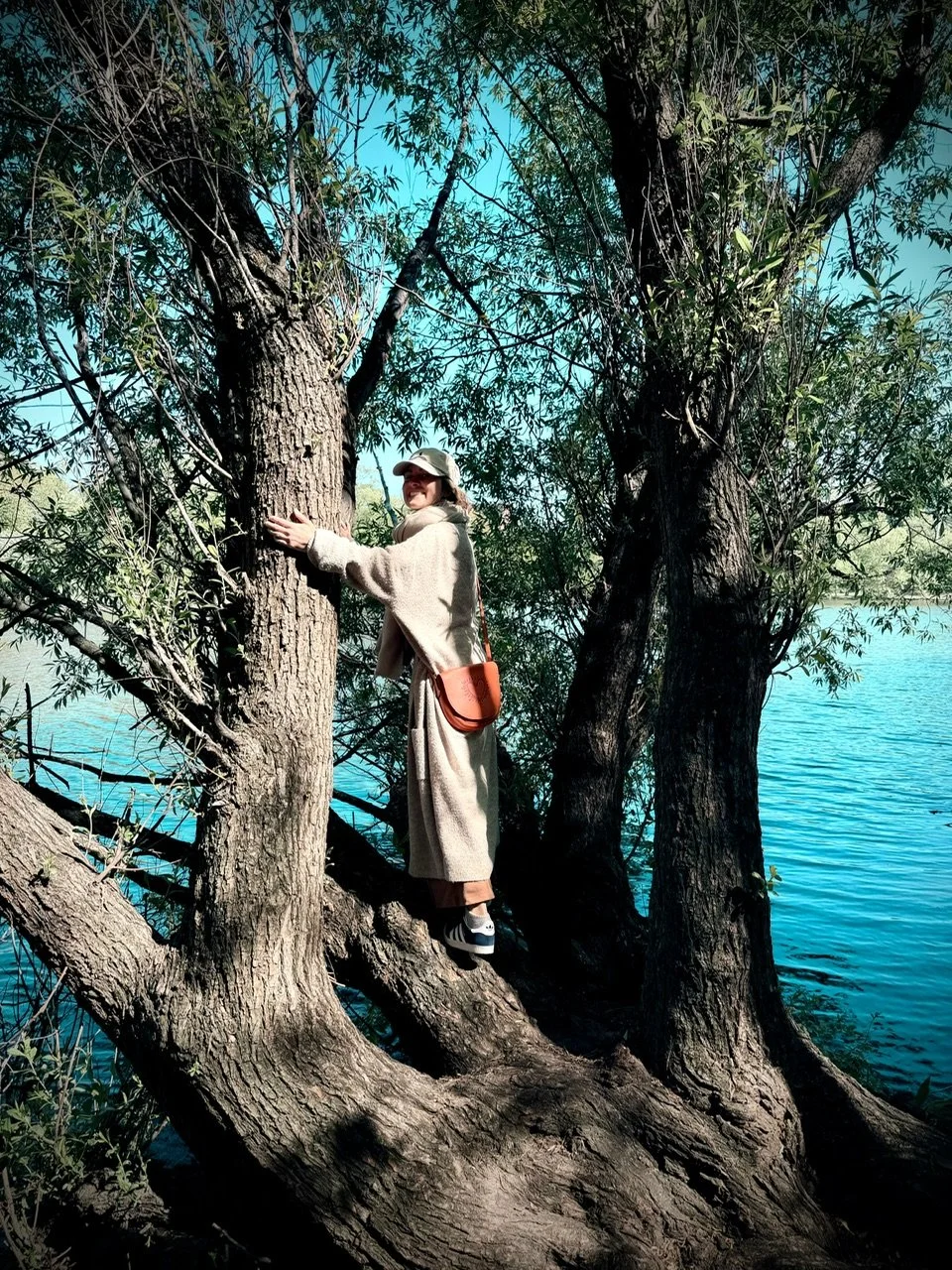Person hugging a tree on a riverbank, wearing a beige coat, hat, and sneakers, with a brown shoulder bag, under a large leafy tree with blue sky and water in the background.