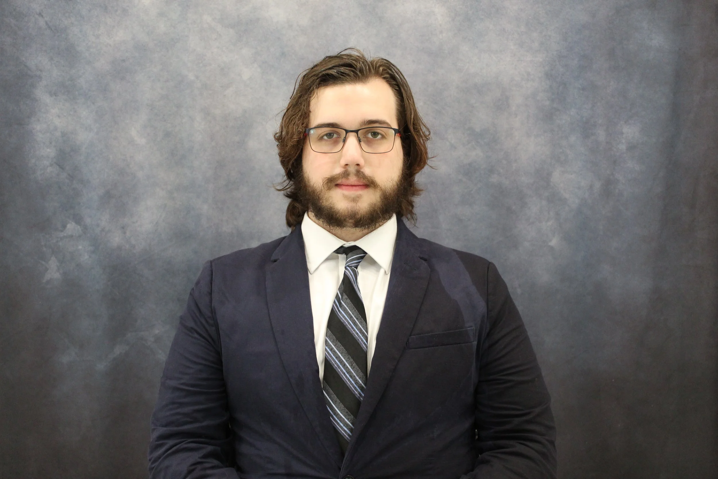 A man with shoulder-length brown hair, glasses, and a beard wearing a suit, white shirt, and striped tie, sitting against a gray textured background.