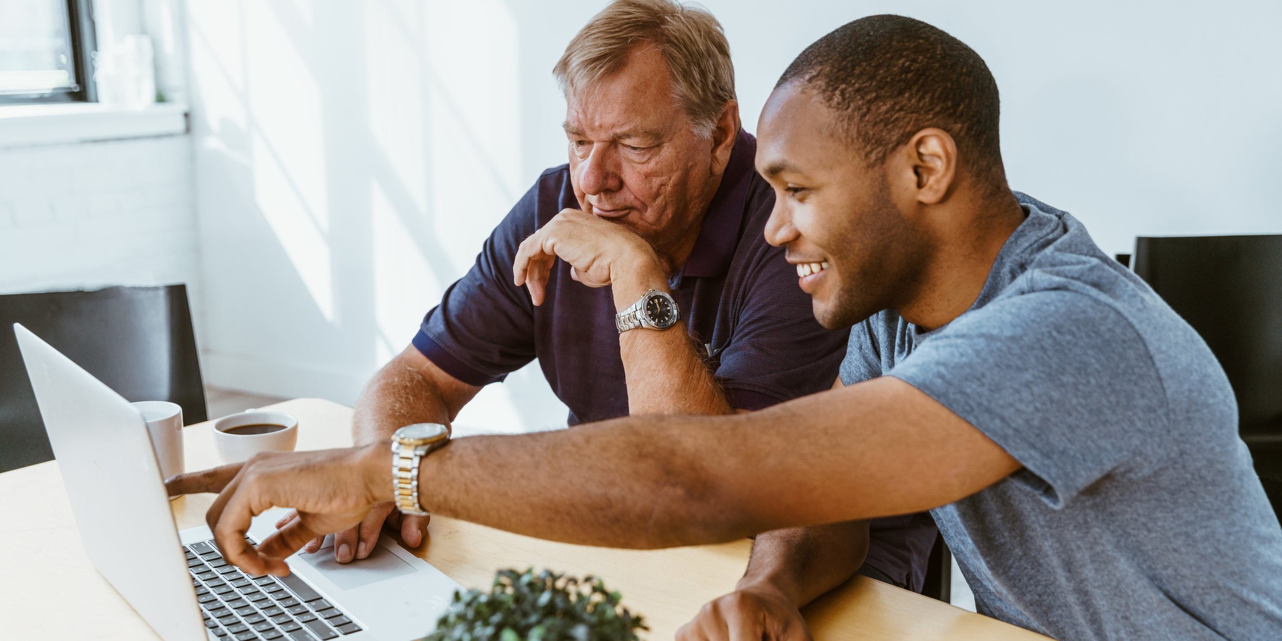 Two men sitting at a desk looking at a laptop, smiling and discussing, with a coffee mug nearby.