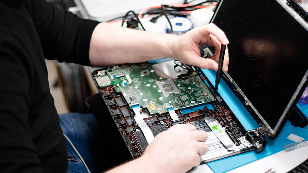 A person repairing a laptop, holding the screen with one hand and using a tool with the other to work on the device's internals, with the laptop open and showing its circuit board and components.