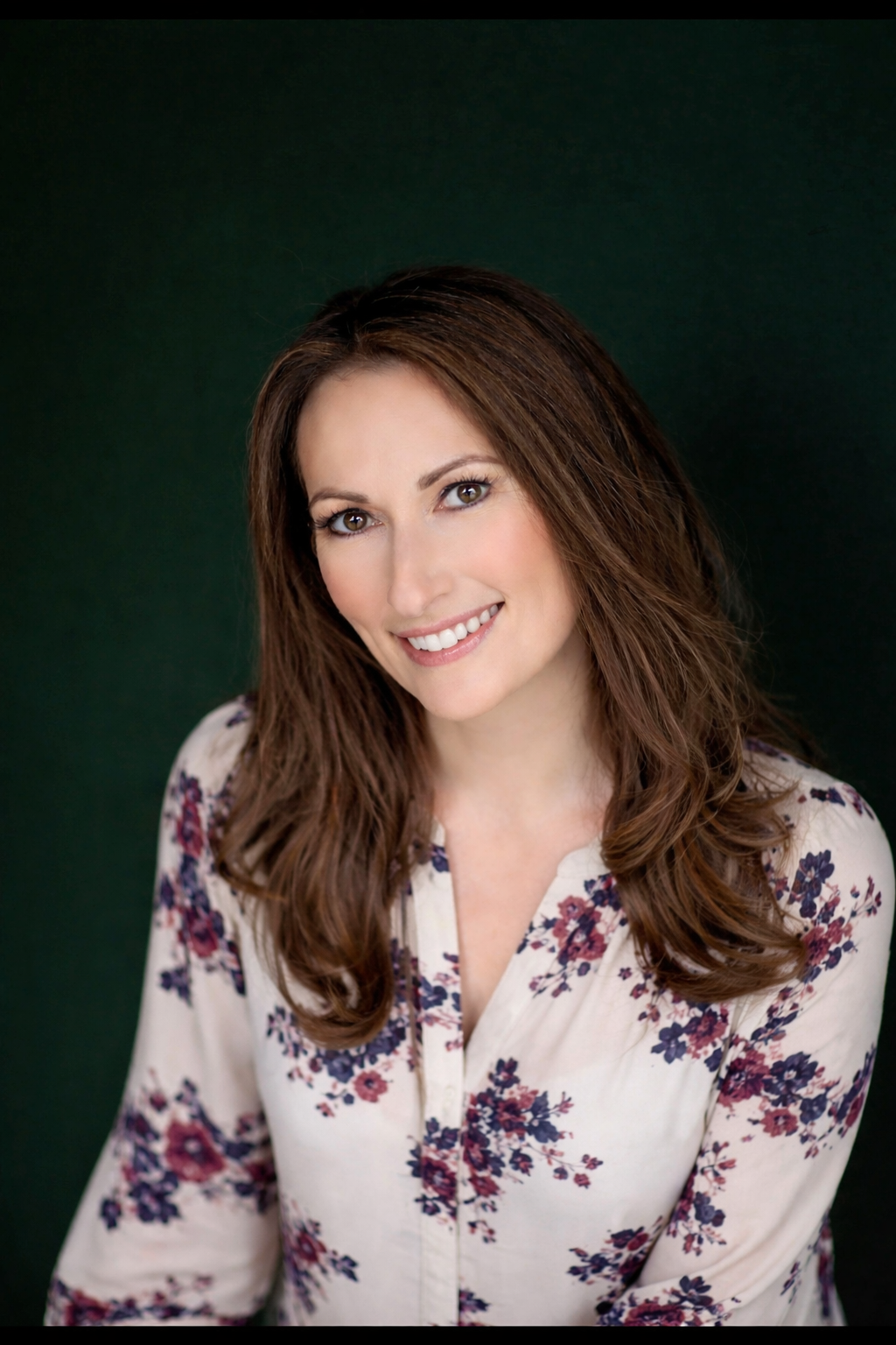 A woman with long, wavy brown hair smiling, wearing a floral blouse against a dark green background.