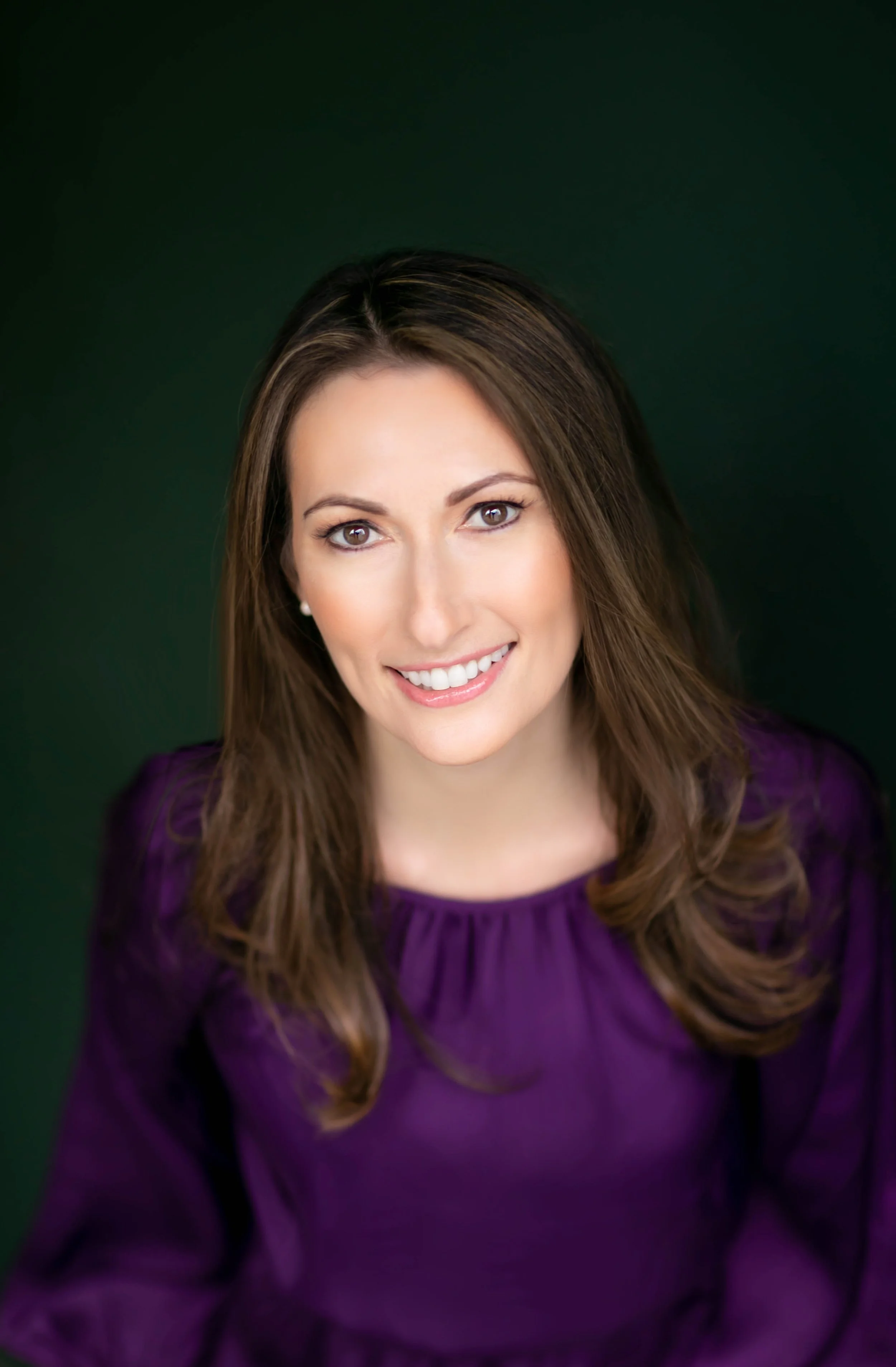 Portrait of a smiling woman with brown hair, wearing a purple top, against a dark green background.