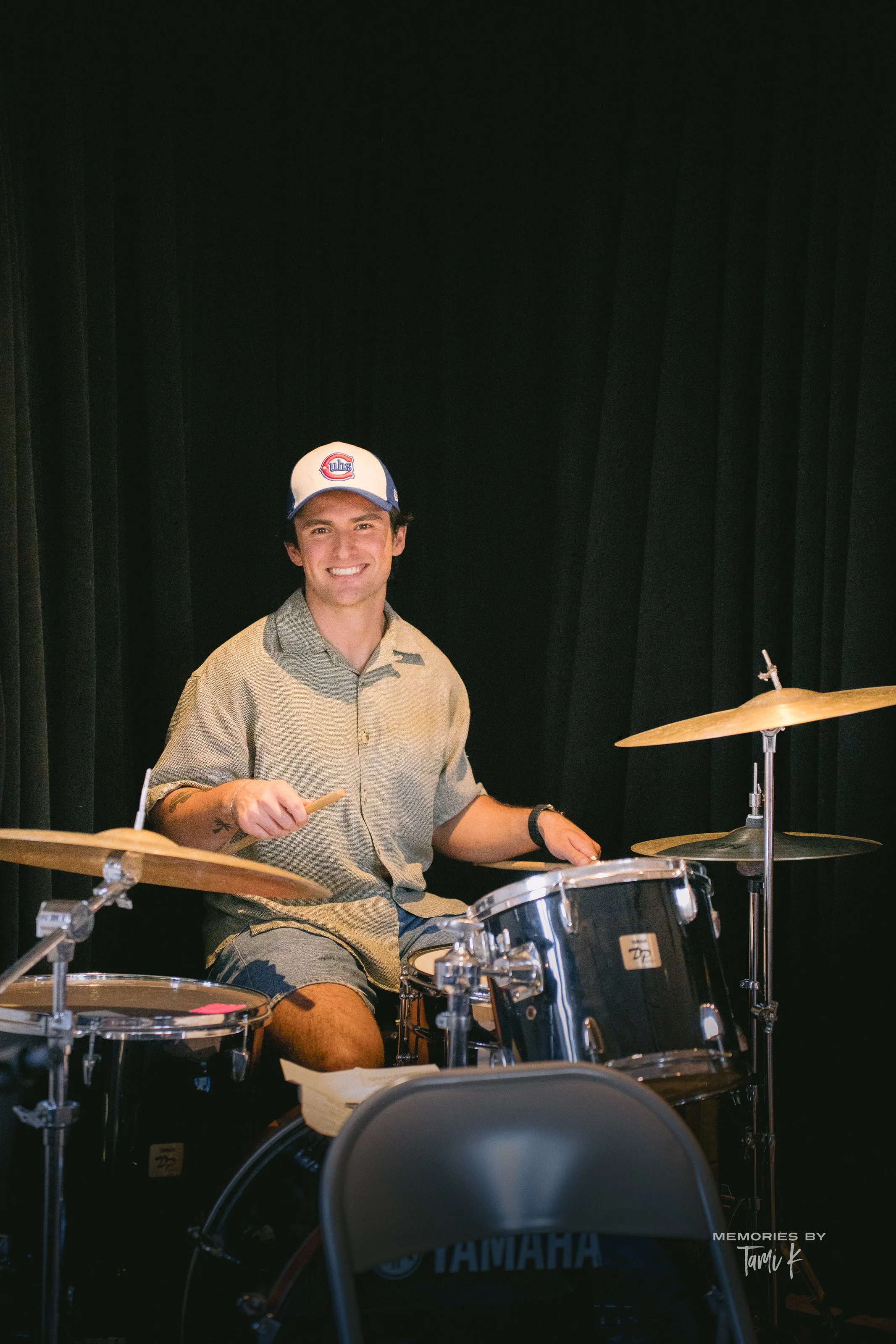 A young man with short dark hair and a bright smile, sitting at a drum set with a black background. He wears a blue and white cap with a logo and a beige, short-sleeved button-up shirt. He is holding drumsticks, and a cymbal and drum are visible in front of him.