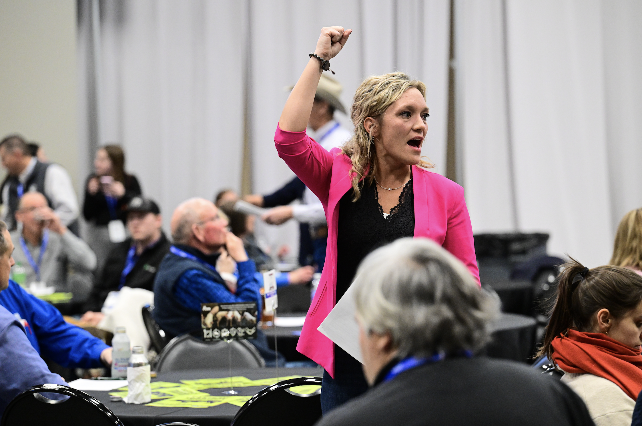 A woman with blonde hair wearing a bright pink blazer raising her fist in the air in a conference room filled with seated attendees listening to her speech.