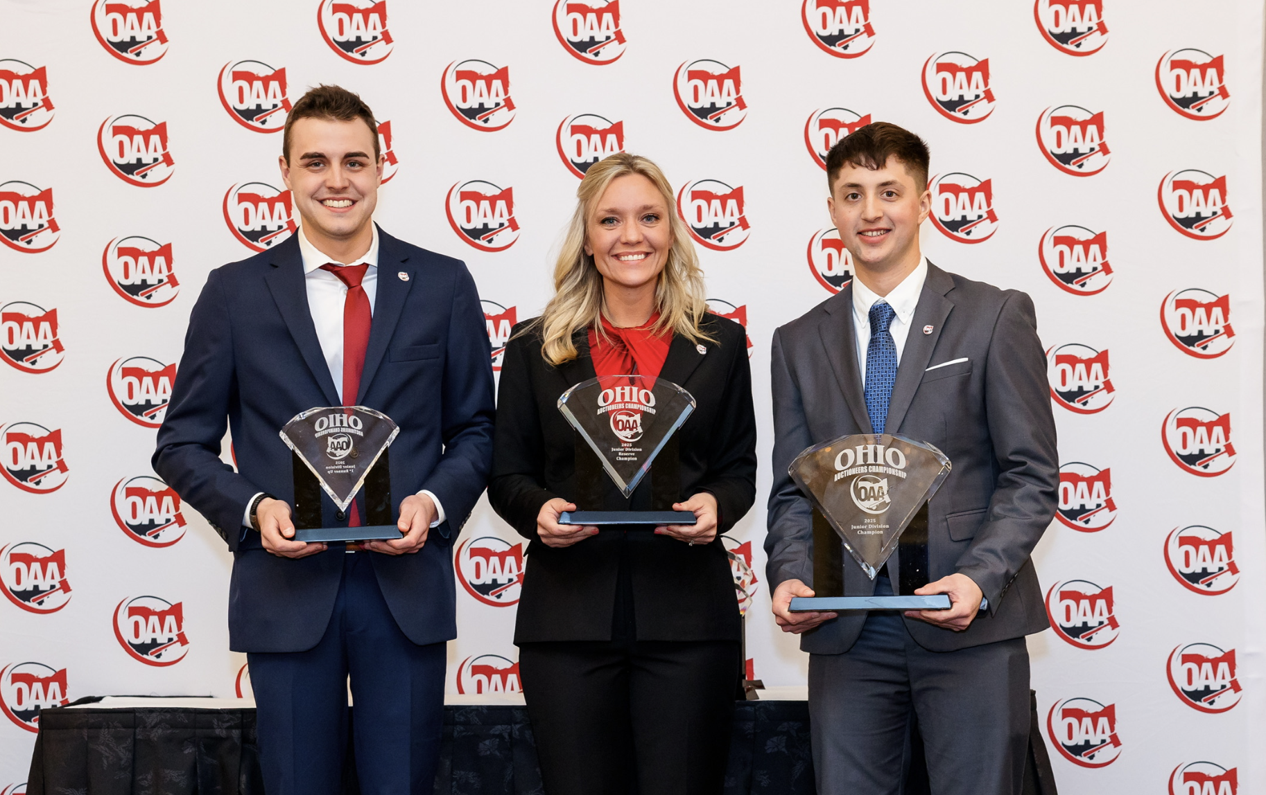 Three people dressed in formal attire holding glass awards during an awards ceremony with a backdrop featuring the logo of Ohio Auctioneers Association (OAA).