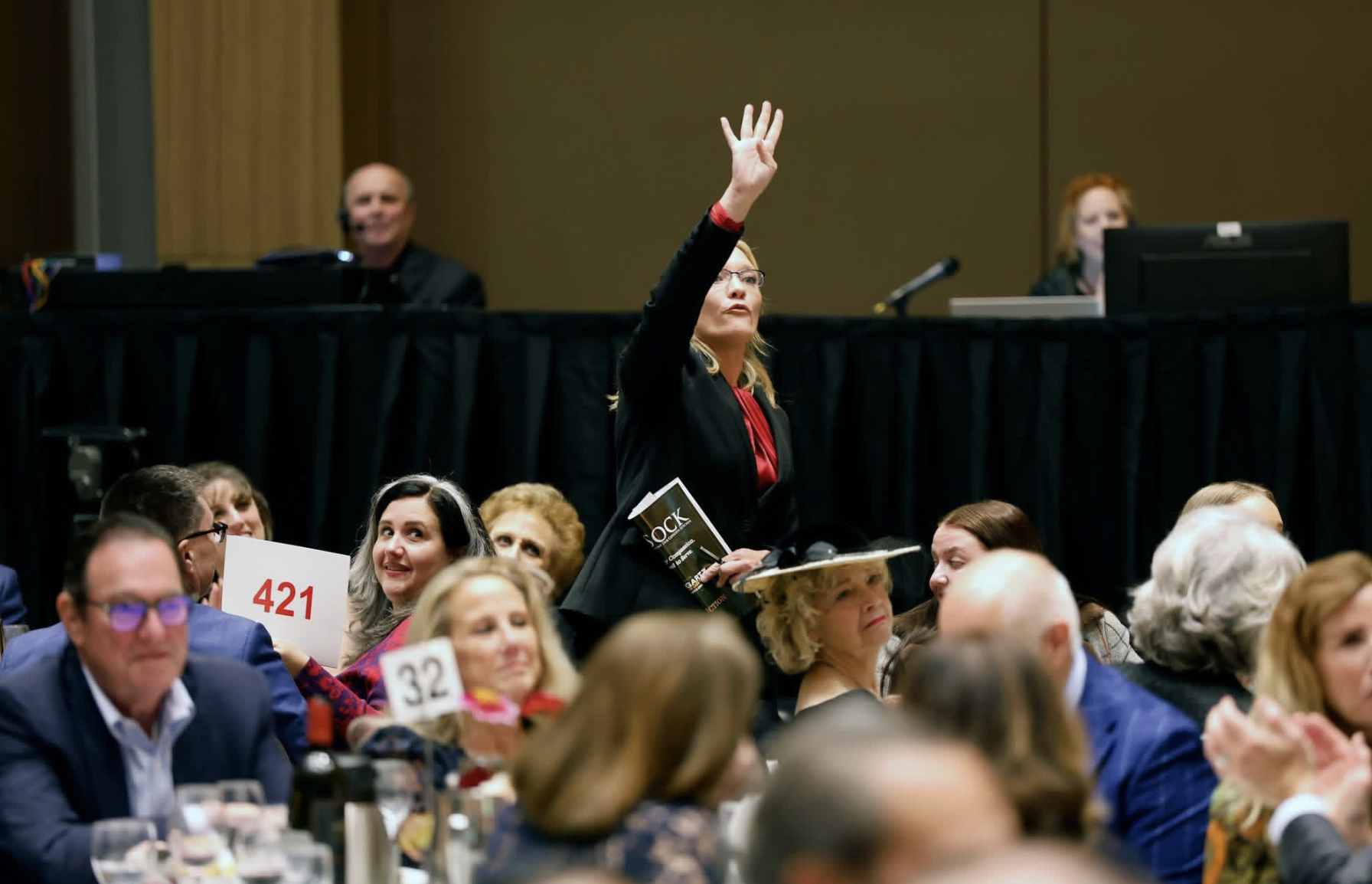 A woman with glasses and blonde hair raising her hand in a crowded conference hall, holding a booklet, surrounded by seated attendees, some holding numbered signs.