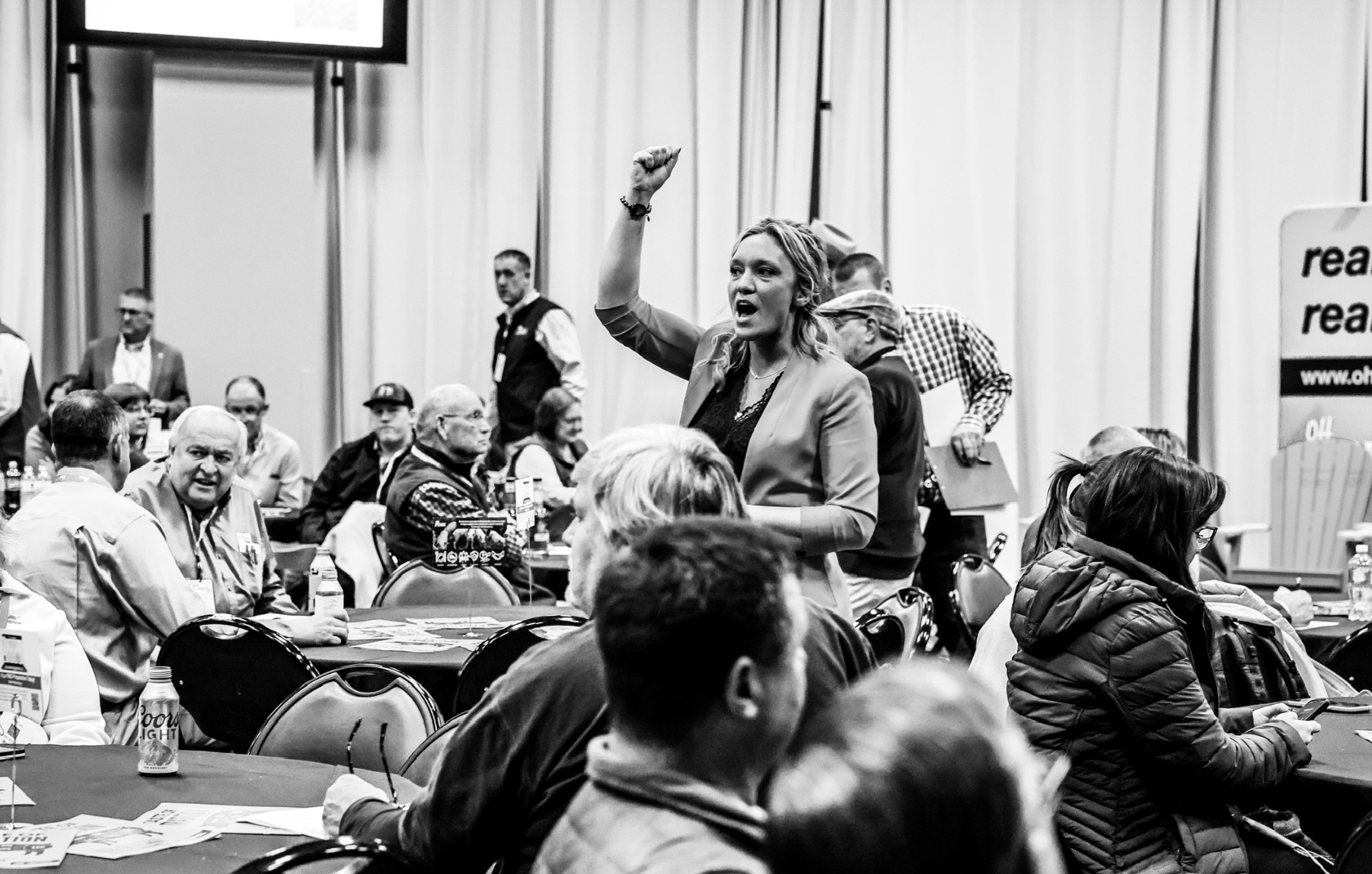 A woman with wavy hair raising her fist in the air while speaking at a crowded indoor event, surrounded by seated attendees and a few standing individuals, some with papers or laptops.