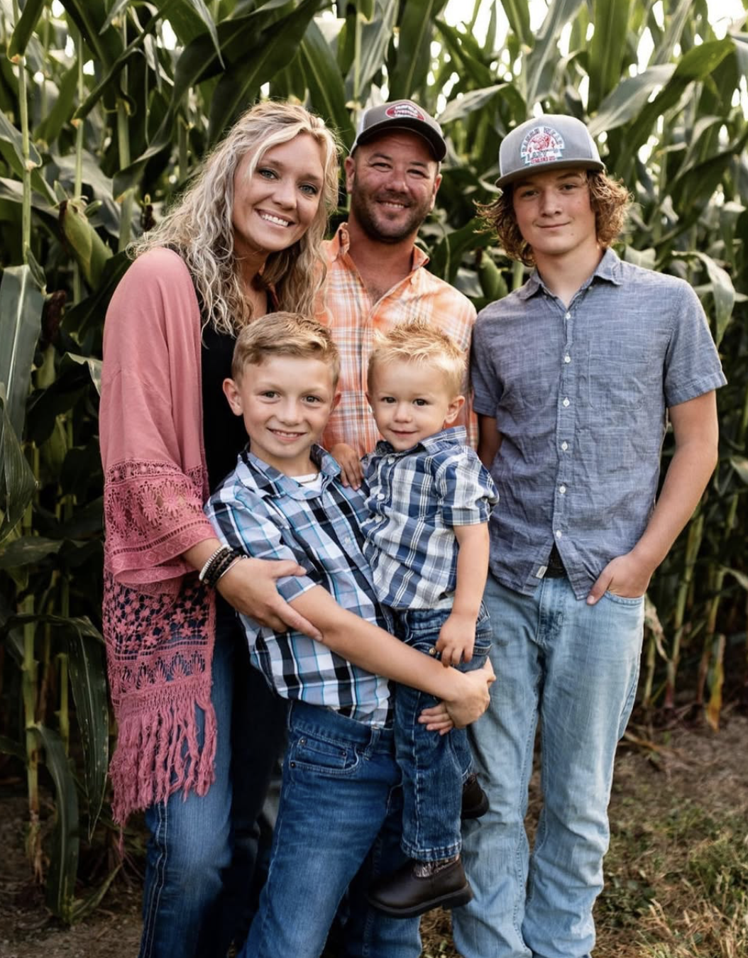 A family of six standing in a cornfield, smiling at the camera. The woman is wearing a pink shawl, the man has a plaid shirt, two boys are wearing plaid shirts, and a teenage boy is in a gray shirt and cap.
