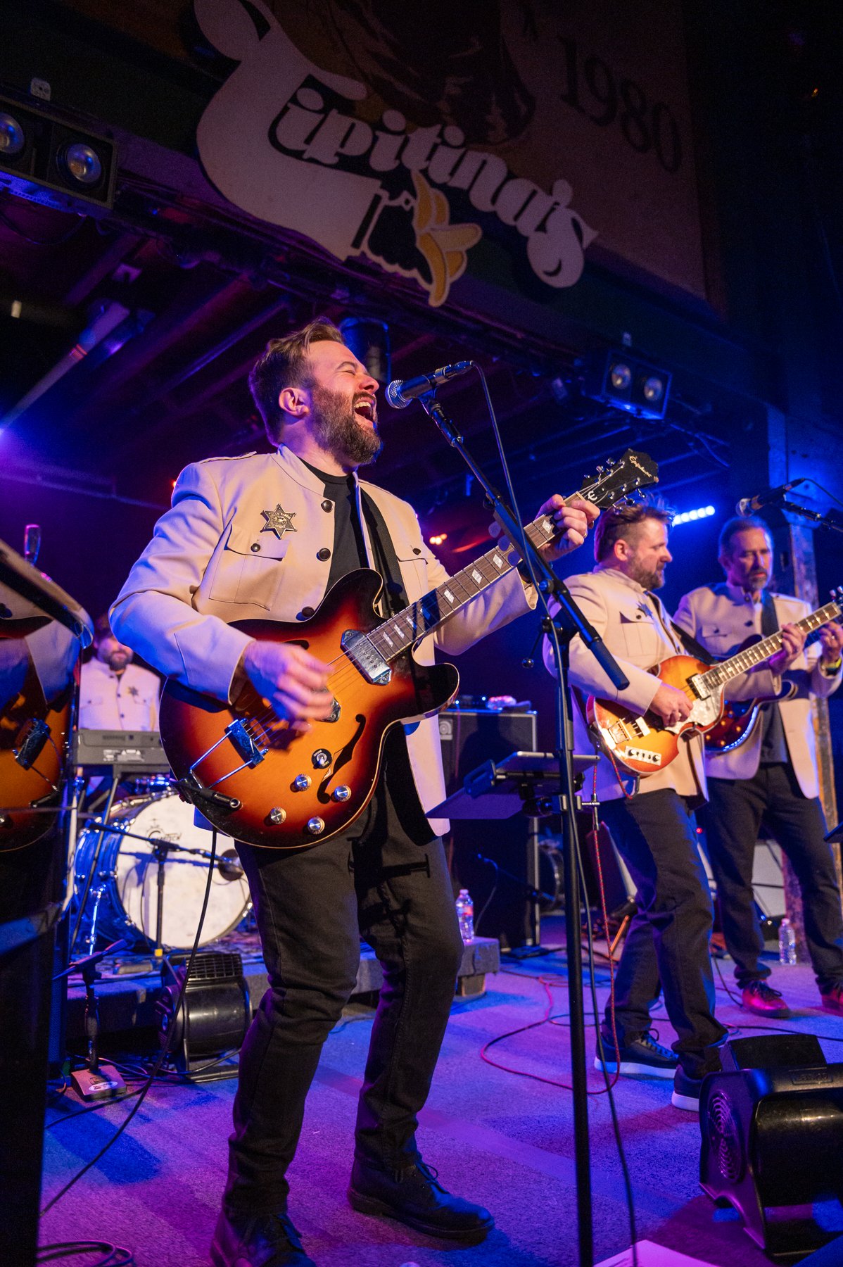 A band performs on stage at Totos, with three members playing guitars and singing. The lead singer is in the foreground, passionately singing with a guitar, wearing a cream-colored jacket with a sheriff badge. The background shows purple lighting and