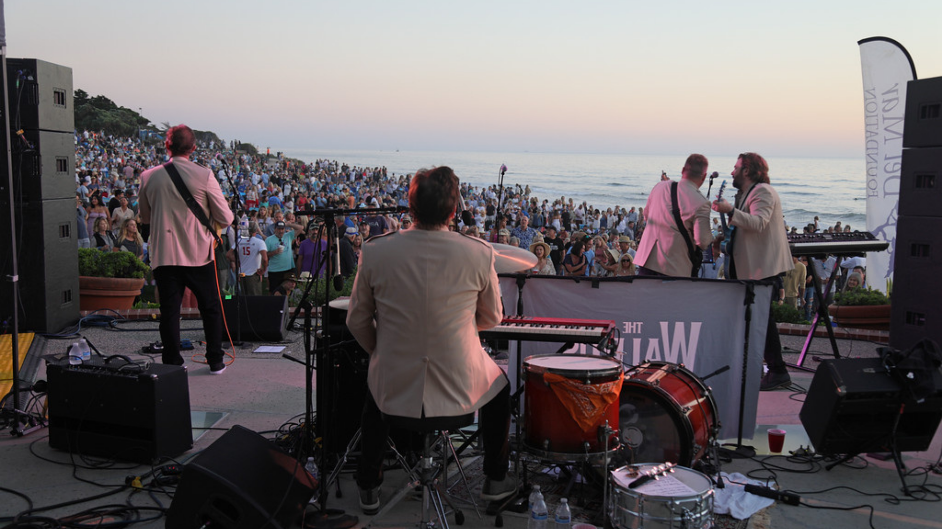 A band performing on stage by the ocean during sunset, with a large audience gathered on the beach.