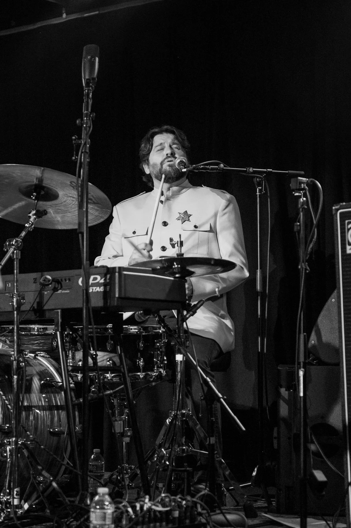 A man with a beard and mustache, wearing a white jacket with a star pin, singing into a microphone while playing drums on stage. Stage equipment such as microphones, drums, a keyboard, and other musical gear are visible.