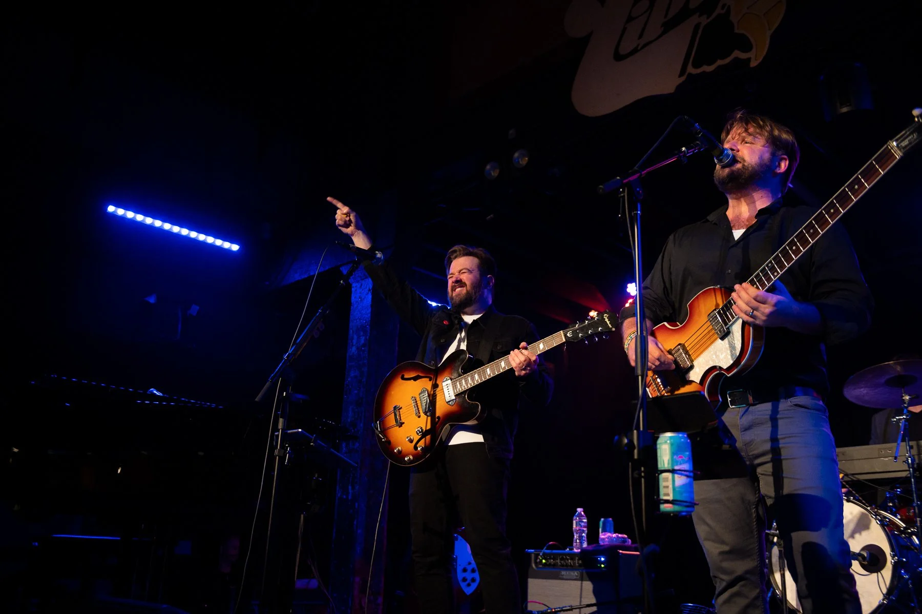 Two male musicians perform on stage with guitars, one singing into a microphone and the other raising a finger, under blue and purple stage lighting.