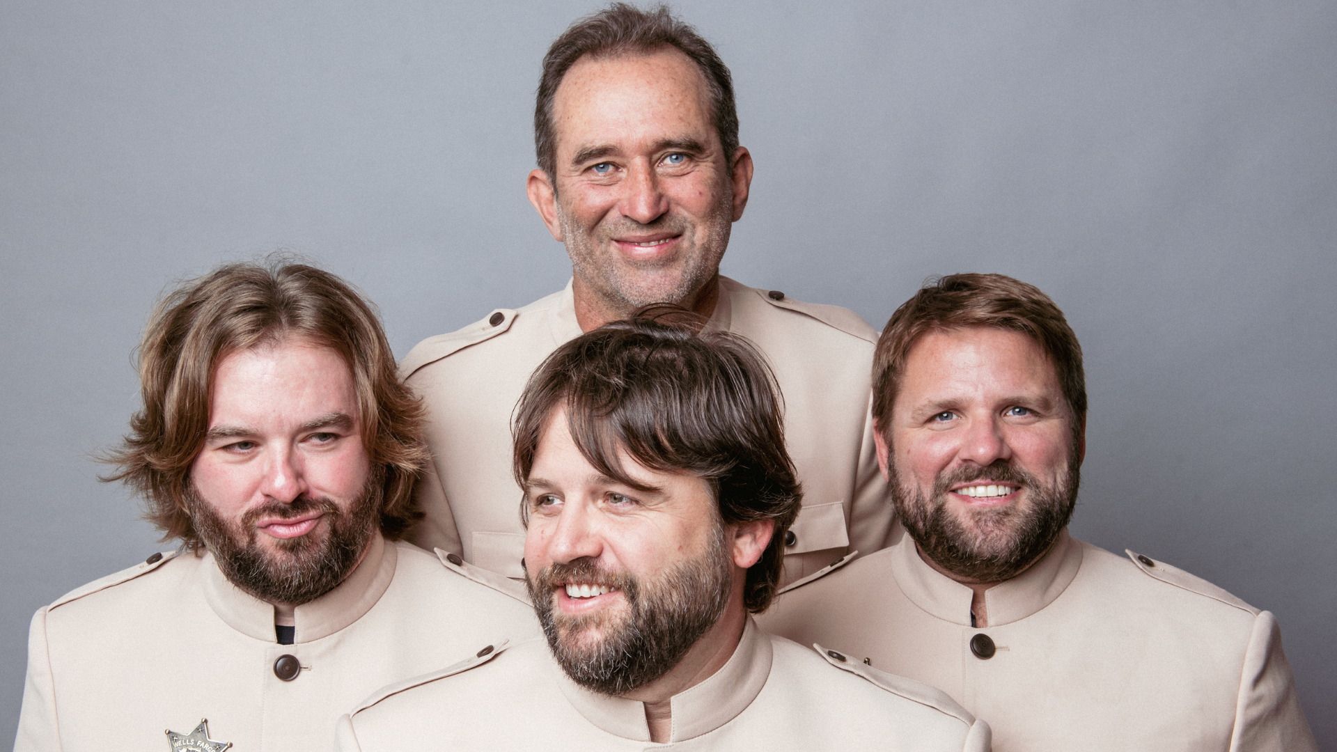 A group photo of five men dressed in beige uniforms, with a grey background.