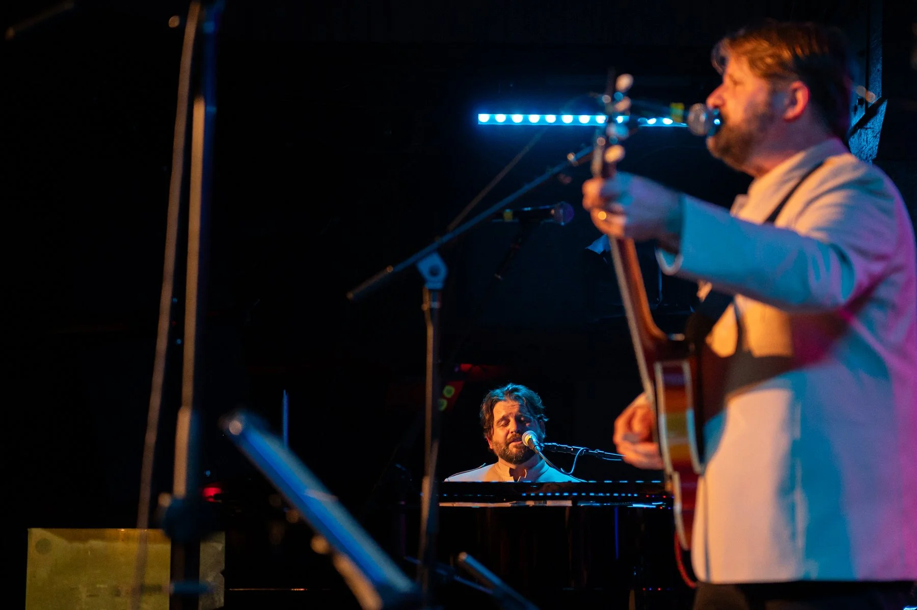 Musicians performing on stage: a man playing guitar and singing, and another man playing piano and singing into microphones, under colorful stage lighting.