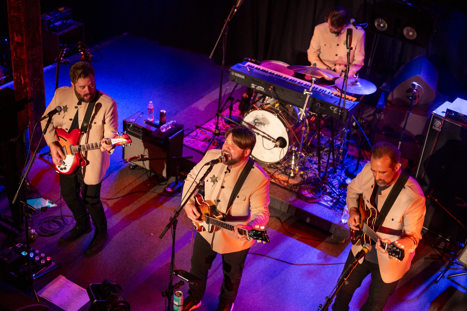 A band performing live on stage with three male singers and two guitarists. The singers are wearing cream-colored jackets and playing guitars, singing into microphones. The keyboardist is playing at a keyboard and a drum set is visible in the backgro