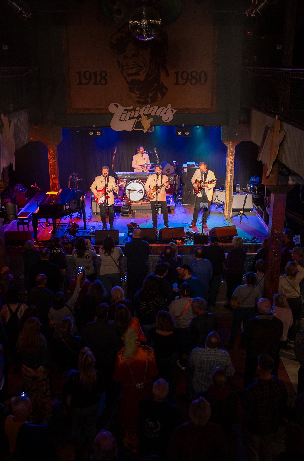 A band performing on stage at a music venue called Tipitina's, with an audience watching. The stage has a black curtain, and four musicians are visible, three playing guitars and one behind a keyboard and drums. The venue's interior features a large 