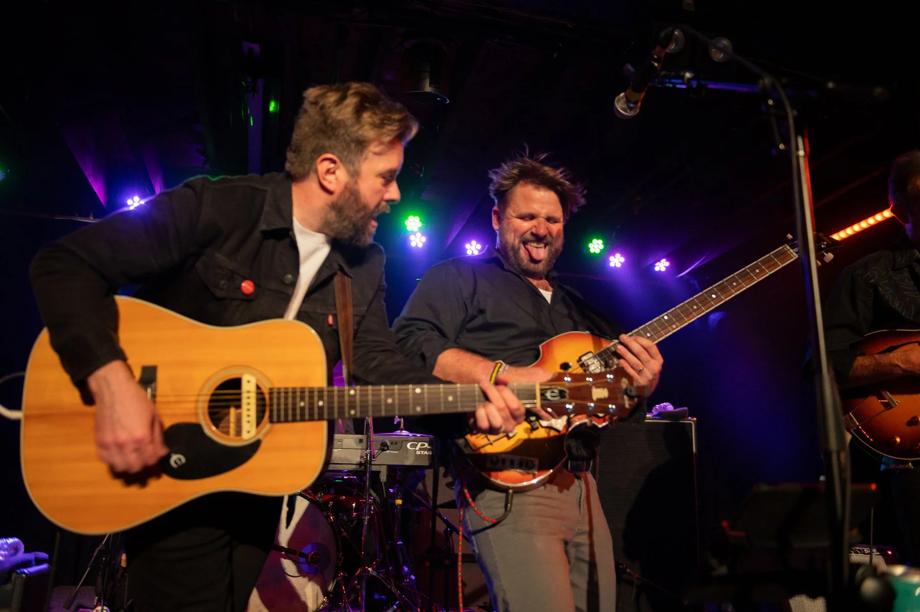 Two men playing guitars on stage with colorful stage lights in the background.