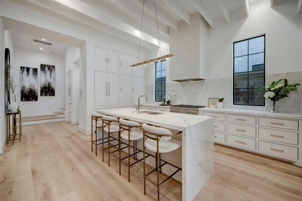 Side view of a luxury white kitchen with an oversized stone island, bar seating and wood flooring in Houston, TX.