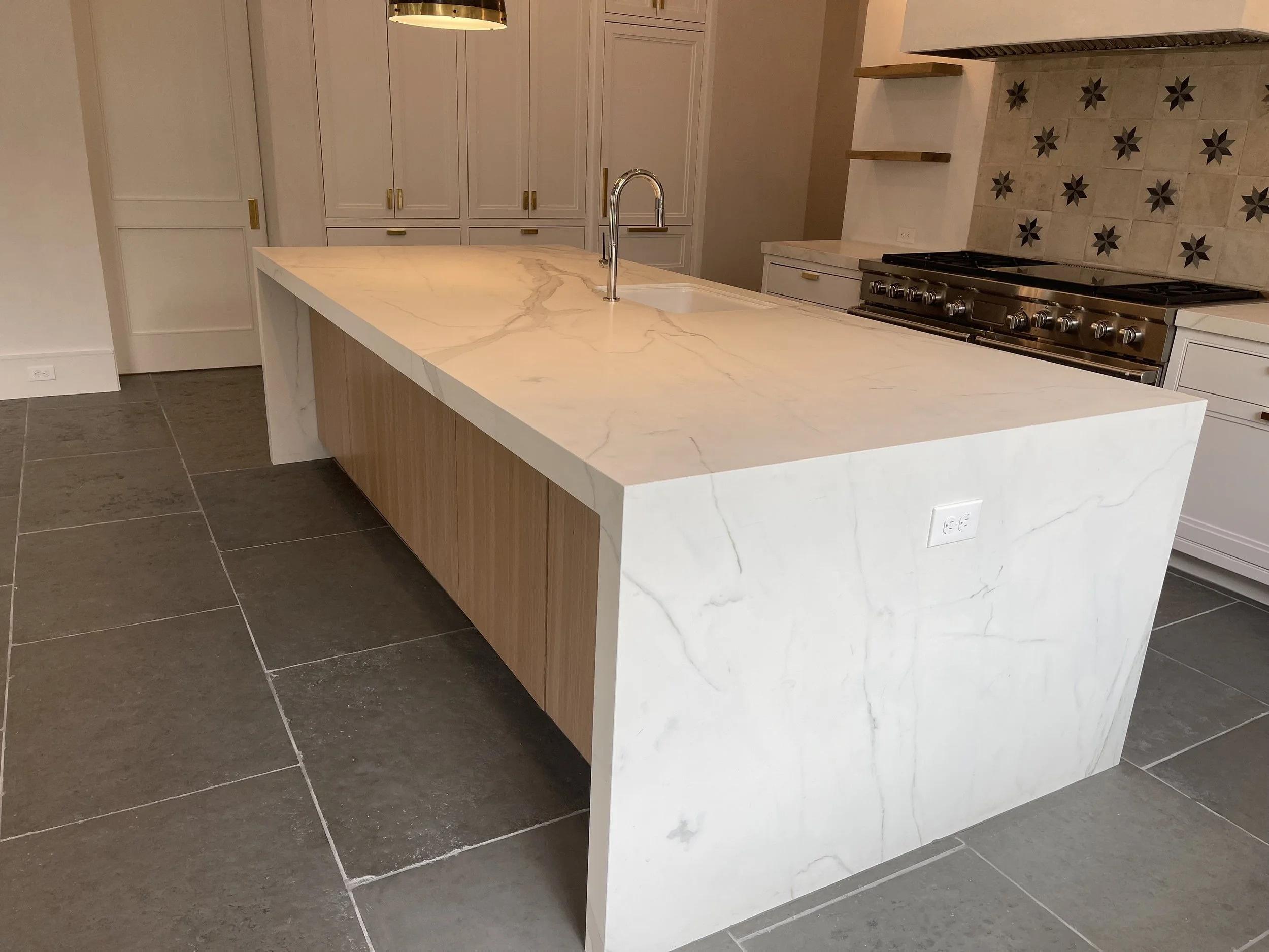 White waterfall-edge stone kitchen island with light wood cabinetry and dark tile flooring in Houston, TX.