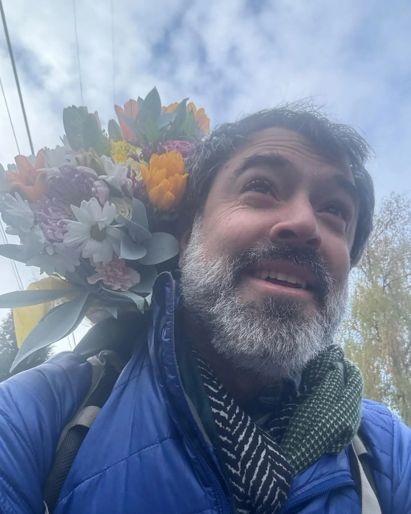 Closeup of man with gray beard carrying flowers in his backpack while riding his bicycle in Portland, OR. Behind him is a blue sky with puffy clouds.