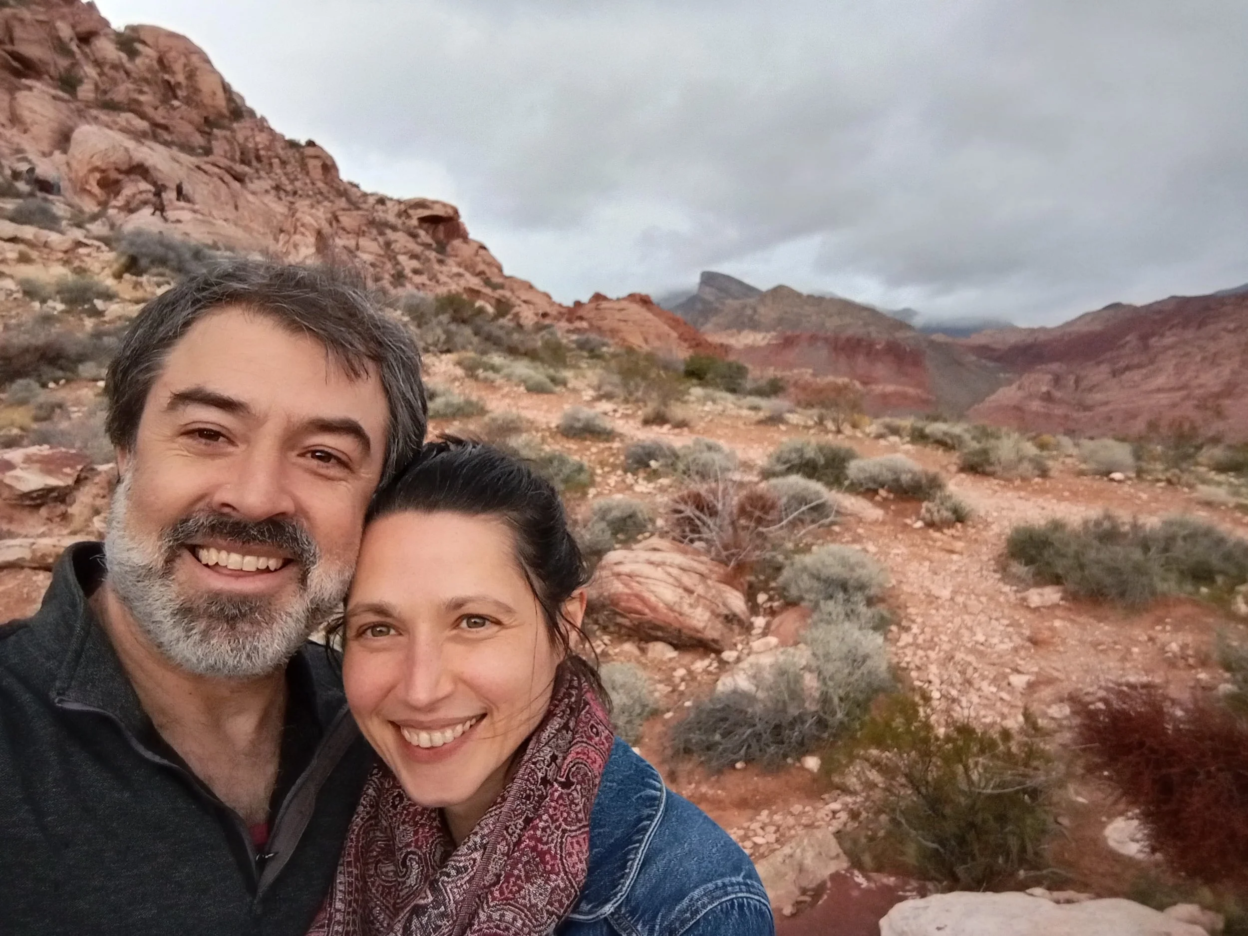 Massage Therapist Eric Fair-Layman (left) and Craniosacral Therapist Erin Boyt (right) face the camera, smiling.  Behind them is a desert landscape with a grayish blue sky.