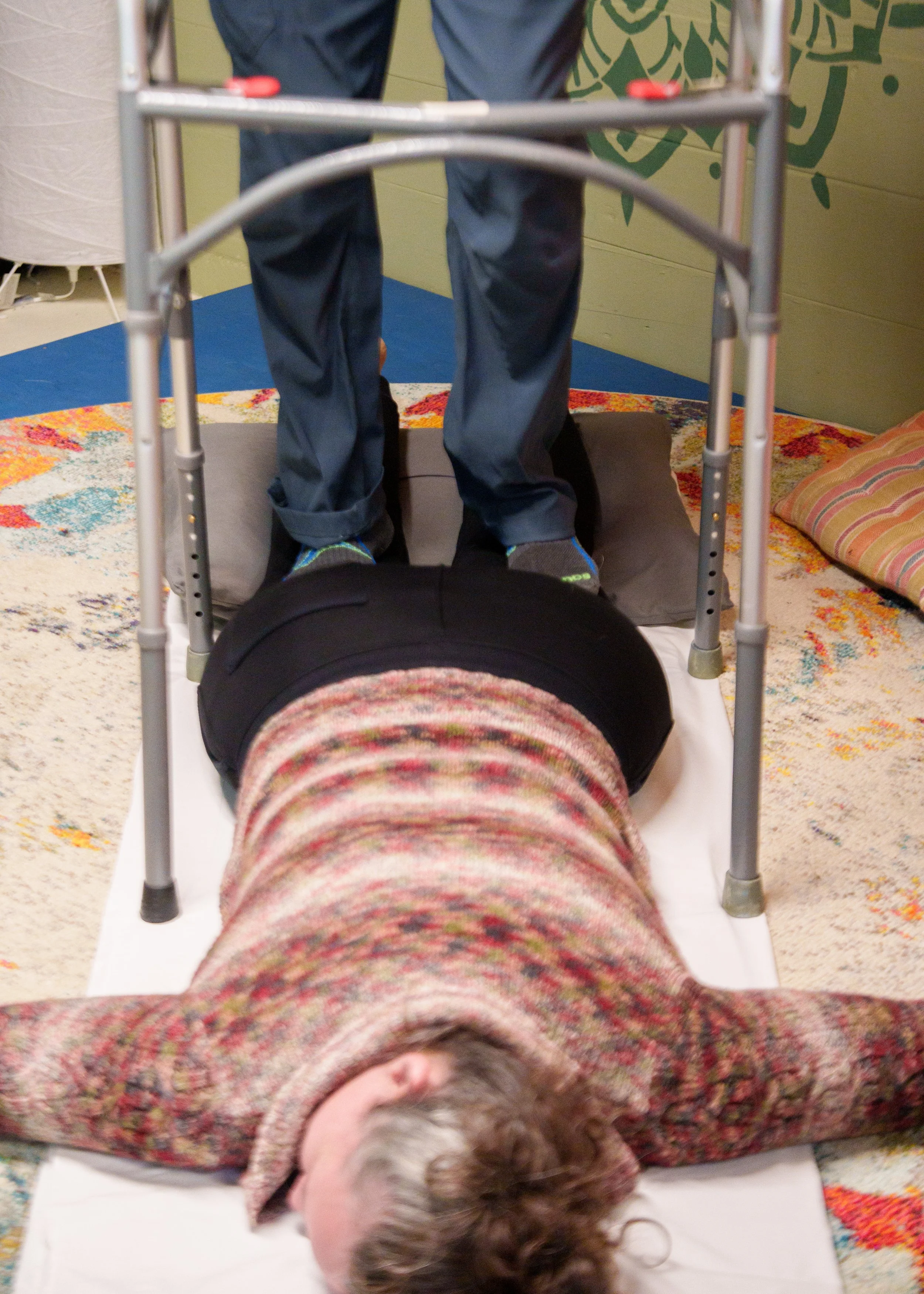 Massage Therapist Eric Fair-Layman walks on the back of a woman's legs. She is laying comfortably on the floor, face down, with her head turned to the right.