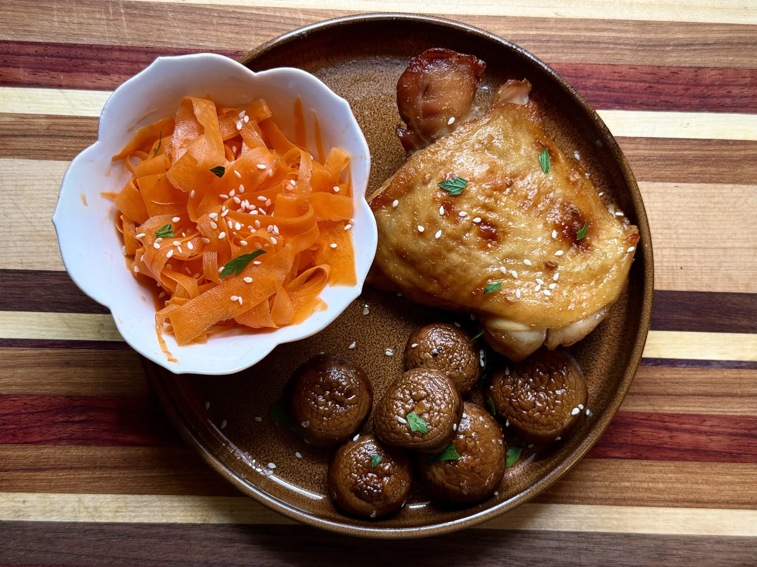 A plate with a serving of baked chicken topped with cheese and green herbs, surrounded by round cooked mushrooms, and a bowl of cooked carrots garnished with sesame seeds and green herbs on a wooden table.
