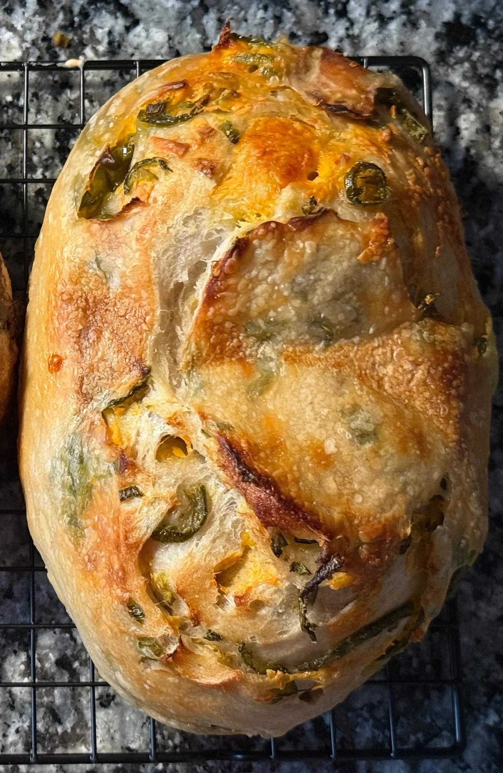 Bread loaf topped with jalapeño slices and melted cheese on a wire rack, placed on a dark granite countertop.