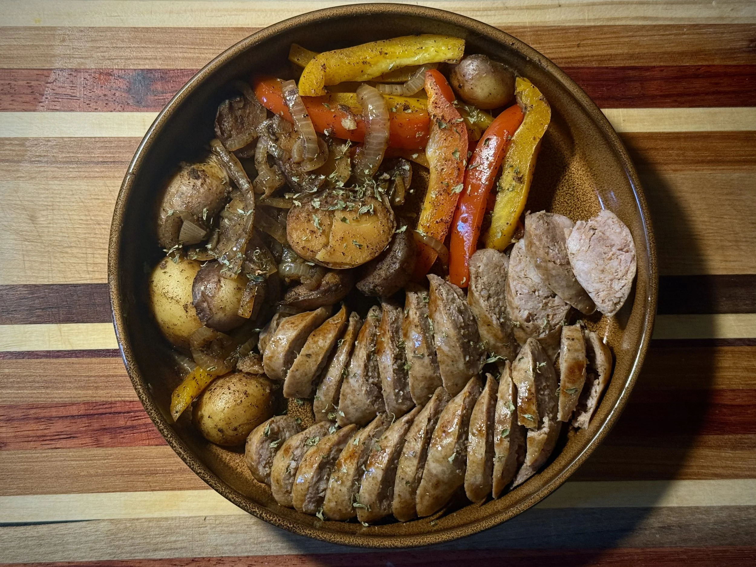 A round dish filled with cooked roasted vegetables including potatoes, carrots, yellow and green peppers, onions, and meat slices, resting on a colorful wooden table.