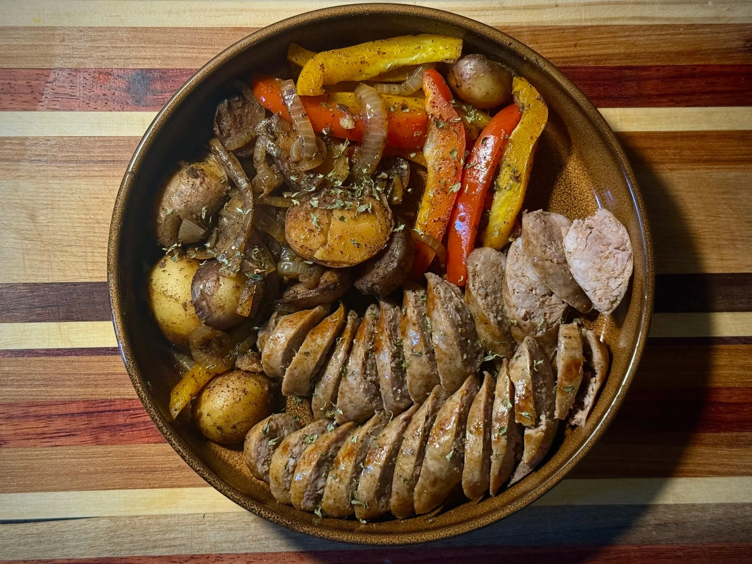 A large bowl of cooked vegetables and meats on a multicolored wooden table, including potatoes, carrots, yellow and green bell peppers, onion, sausage, and slices of grilled meat garnished with herbs.