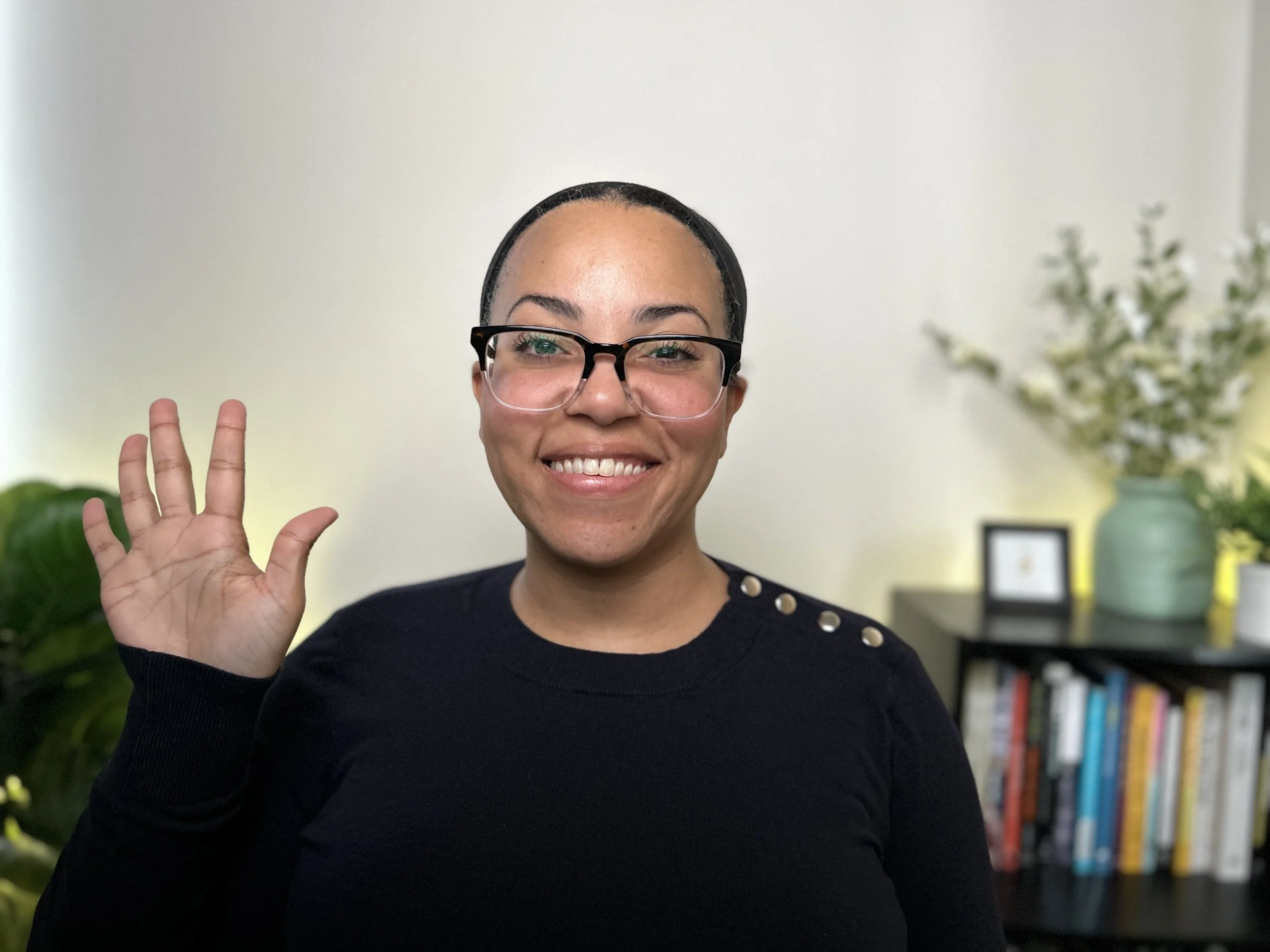 A medium complexion woman with glasses smiling and waving wearing a blue sweater