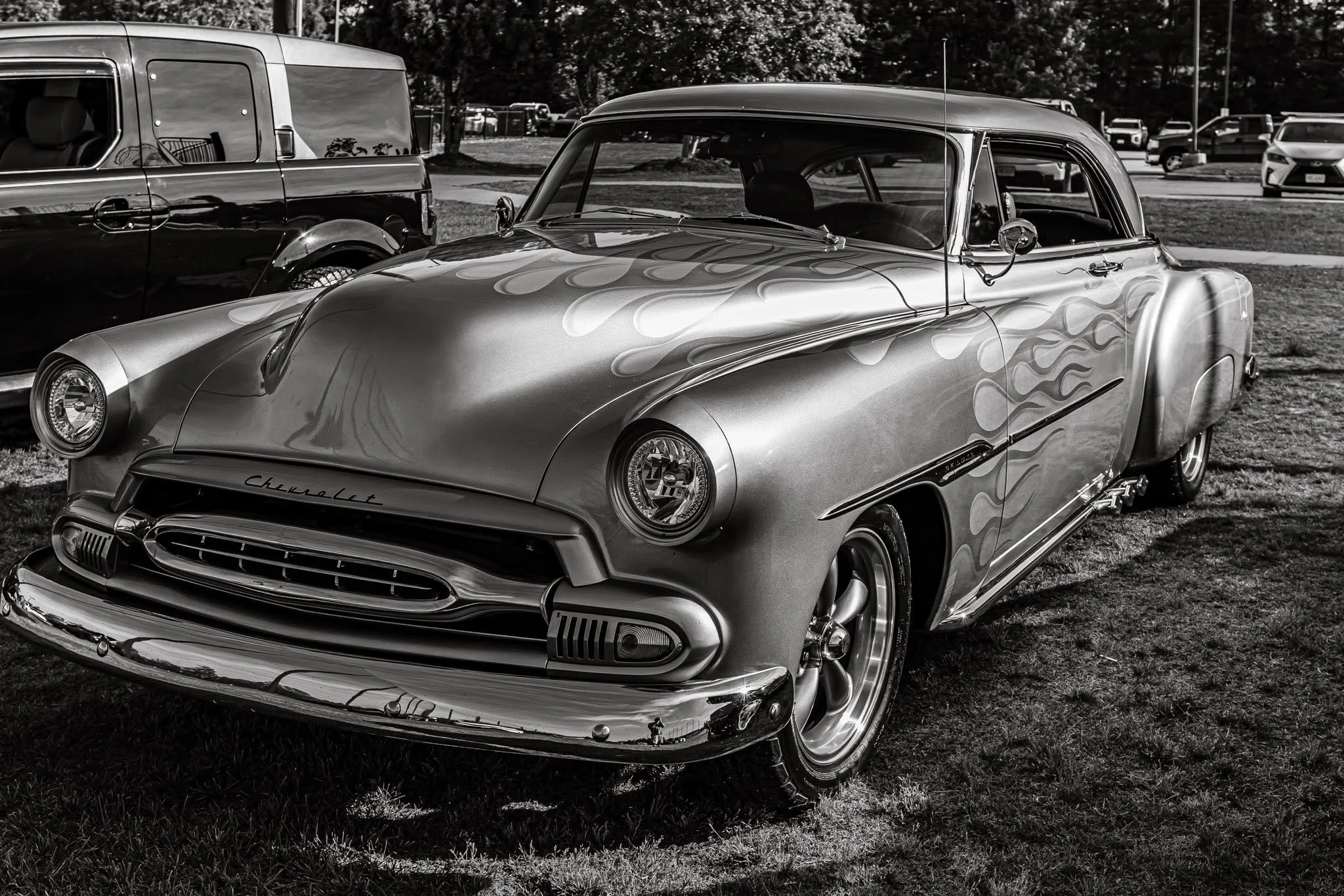 Captured in timeless black and white, a vintage Chevy proudly holds its ground on the show field, its iconic flame decals hinting at a wild past. It’s more than just a car—it’s a rolling piece of history waiting for the next generation of storyteller