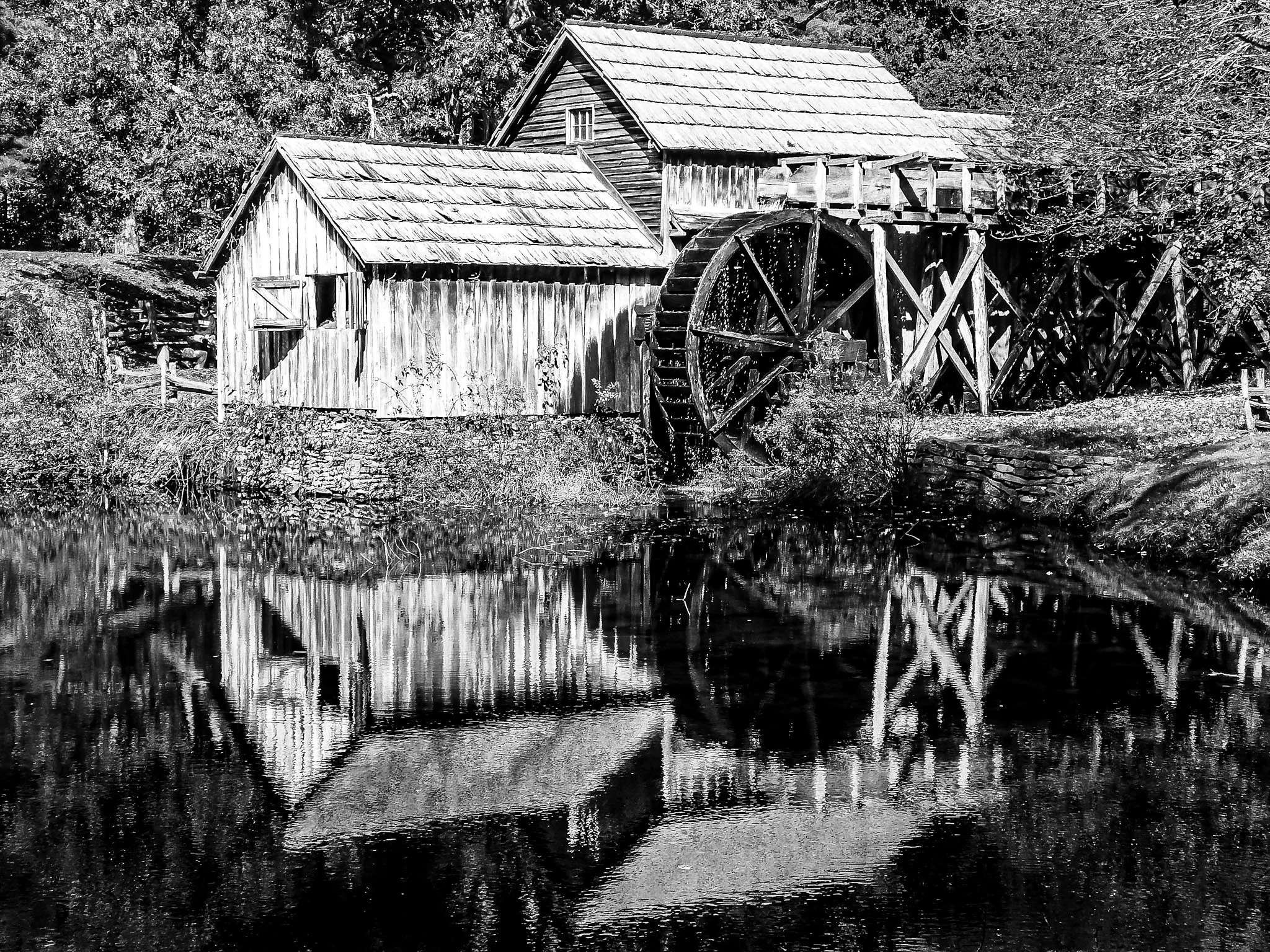 This old black-and-white photograph holds the quiet dignity of early industry. The towering waterwheel rests against the rustic wooden mill. Looking at its reflection rippling in the water below, you can almost hear the creak of the timber and the ru