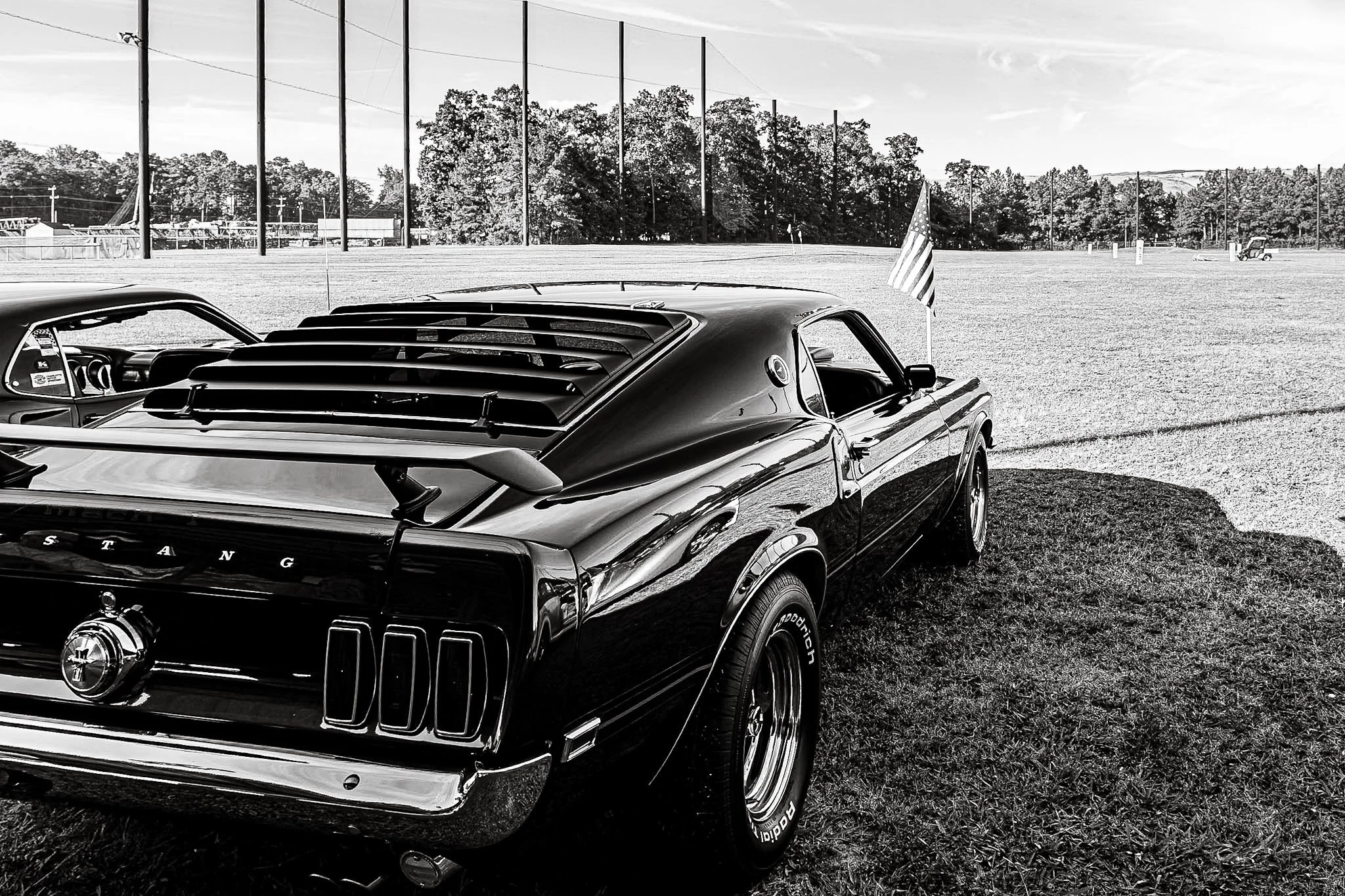 A timeless slice of Americana:  A classic vintage Ford Mustang, sitting beautifully on the show field grass. In the background, Old Glory waves proudly. Captured in a striking, high-contrast black and white photograph that feels like a step back in t