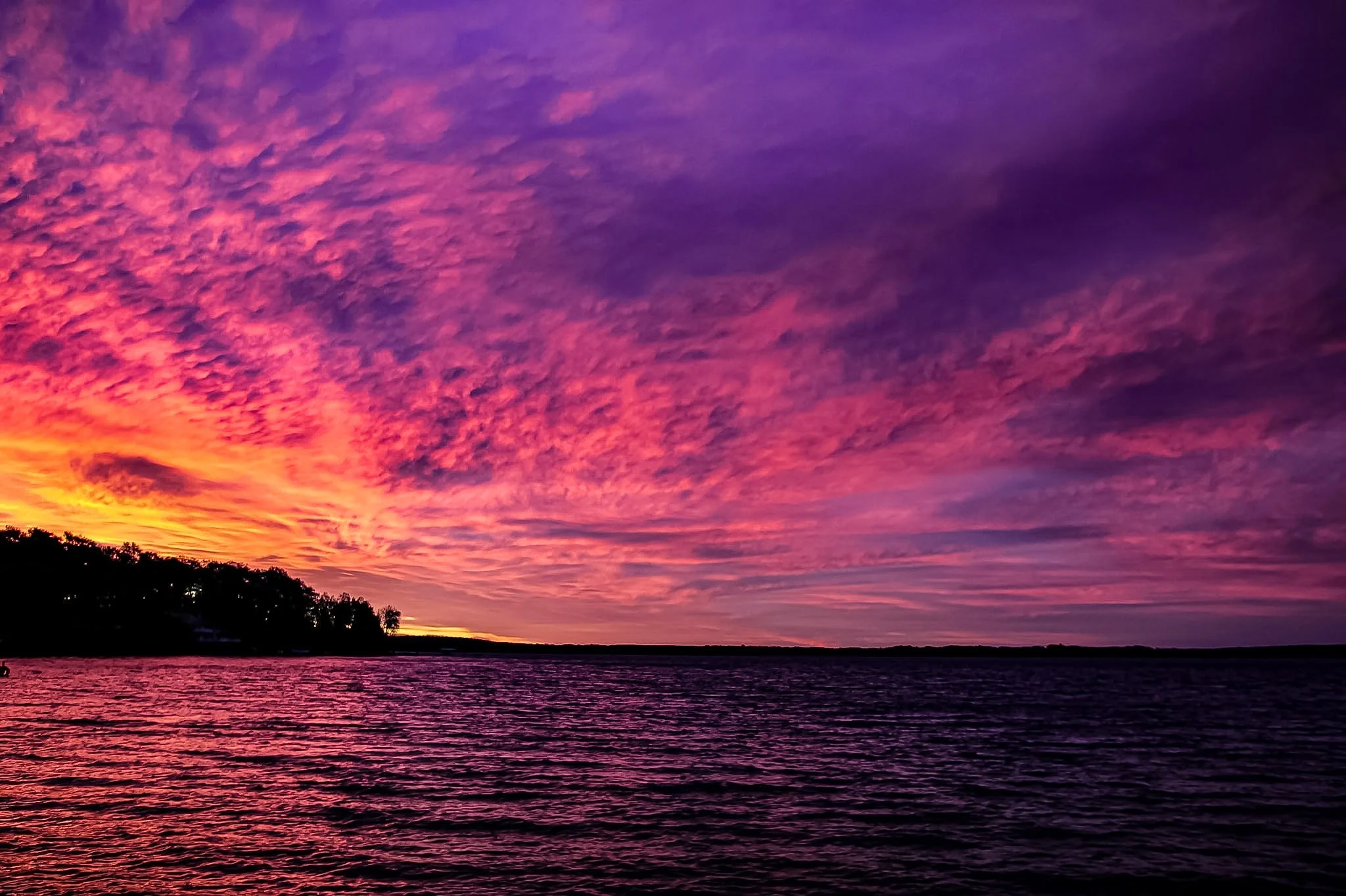 The sky completely showed off tonight—bleeding from a fiery orange into that deep, bruised purple. I just sat there watching the water catch all that color, with the trees framing it like a perfect postcard. Moments like this make everything else qui