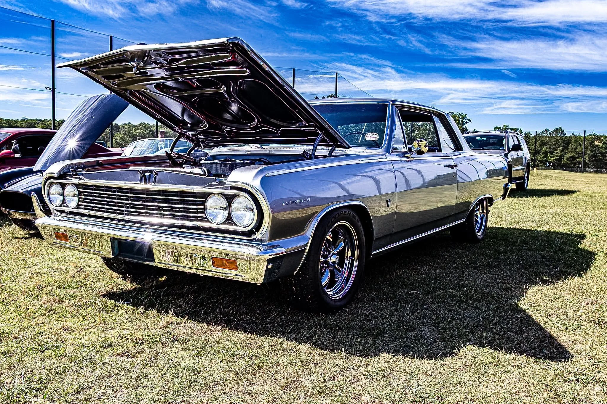 A classic silver Chevrolet, its chrome accents catching the bright afternoon light. With the hood propped open to show off the work under the hood, pure muscle in all its glory.