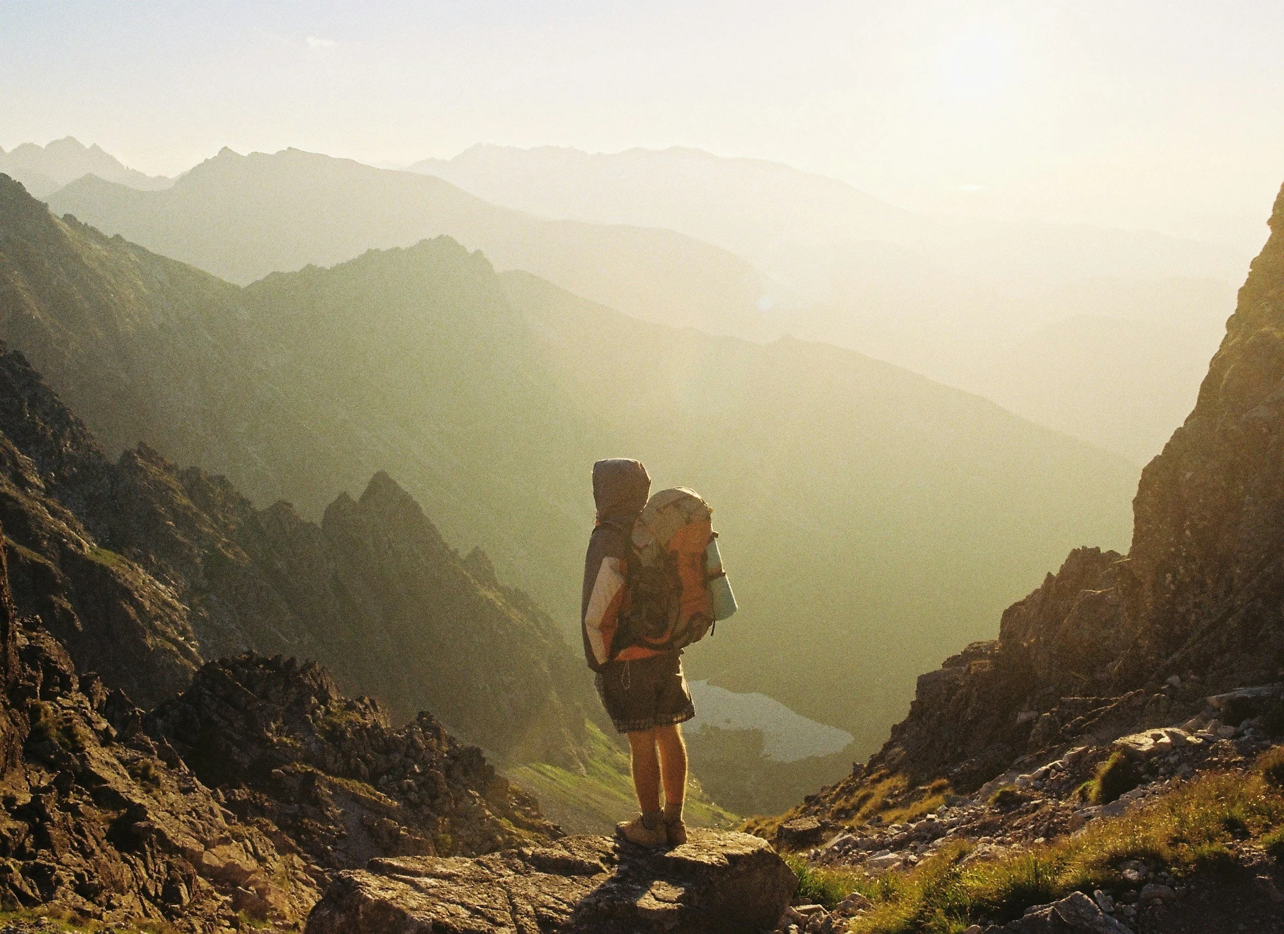 Hiker standing on rocky terrain in a mountainous landscape with multiple mountain ridges in the background, bathed in warm sunlight.