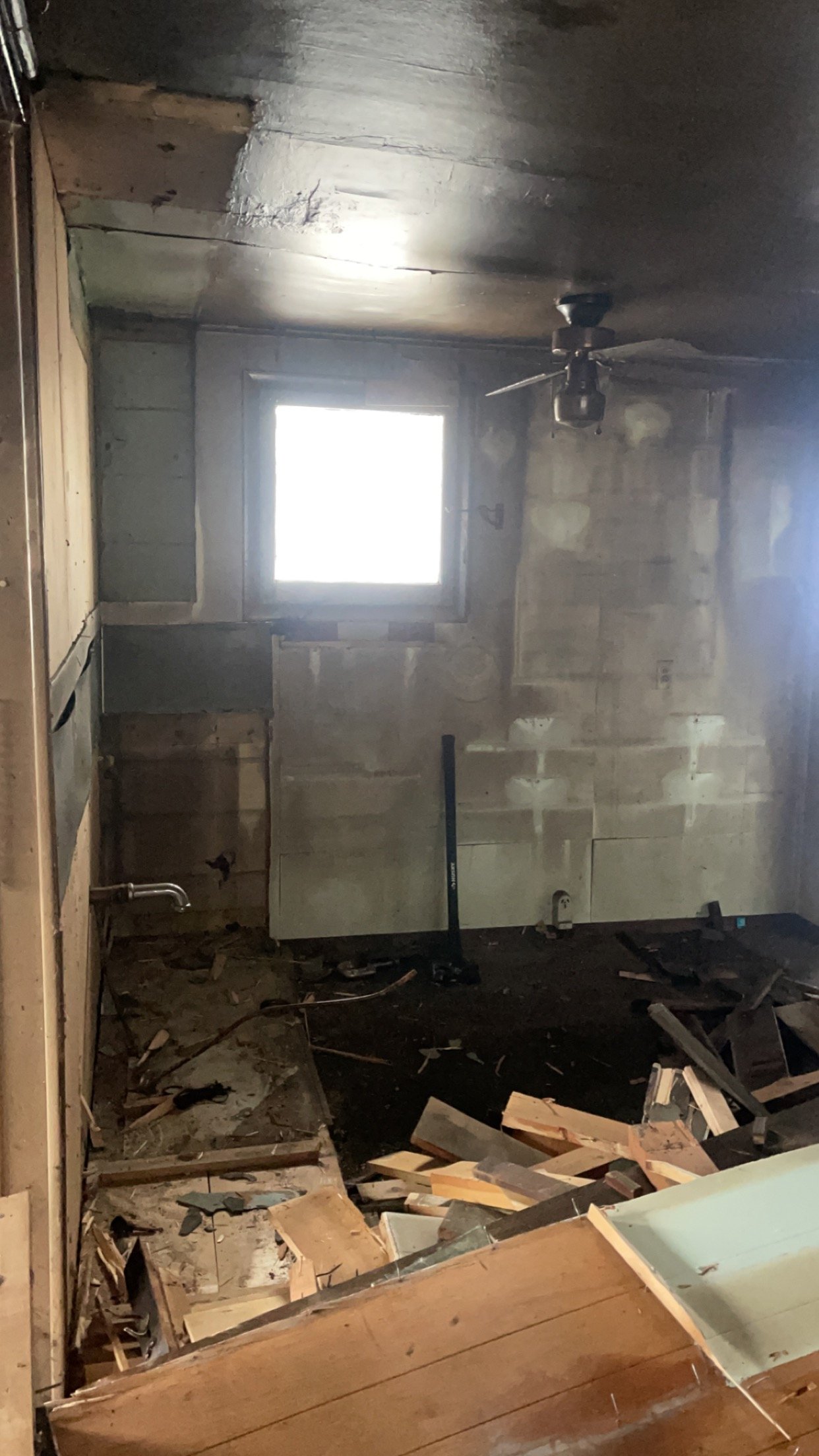 Interior of an unfinished or abandoned room with exposed wooden beams, a single window, scattered debris, and a ceiling fan.