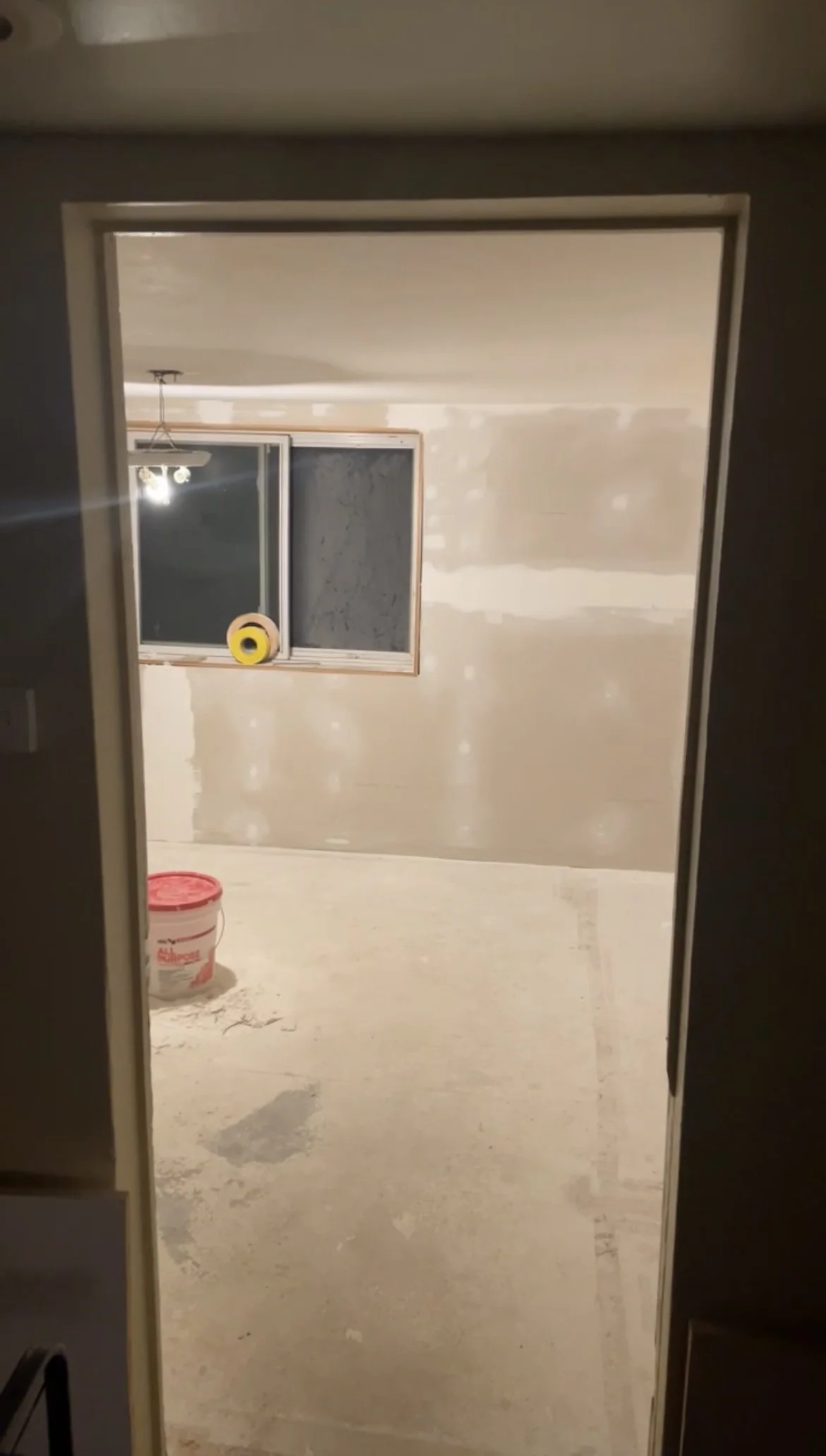 Room under construction with unfinished drywall, a window, a ceiling light fixture, and a bucket of joint compound on the floor.