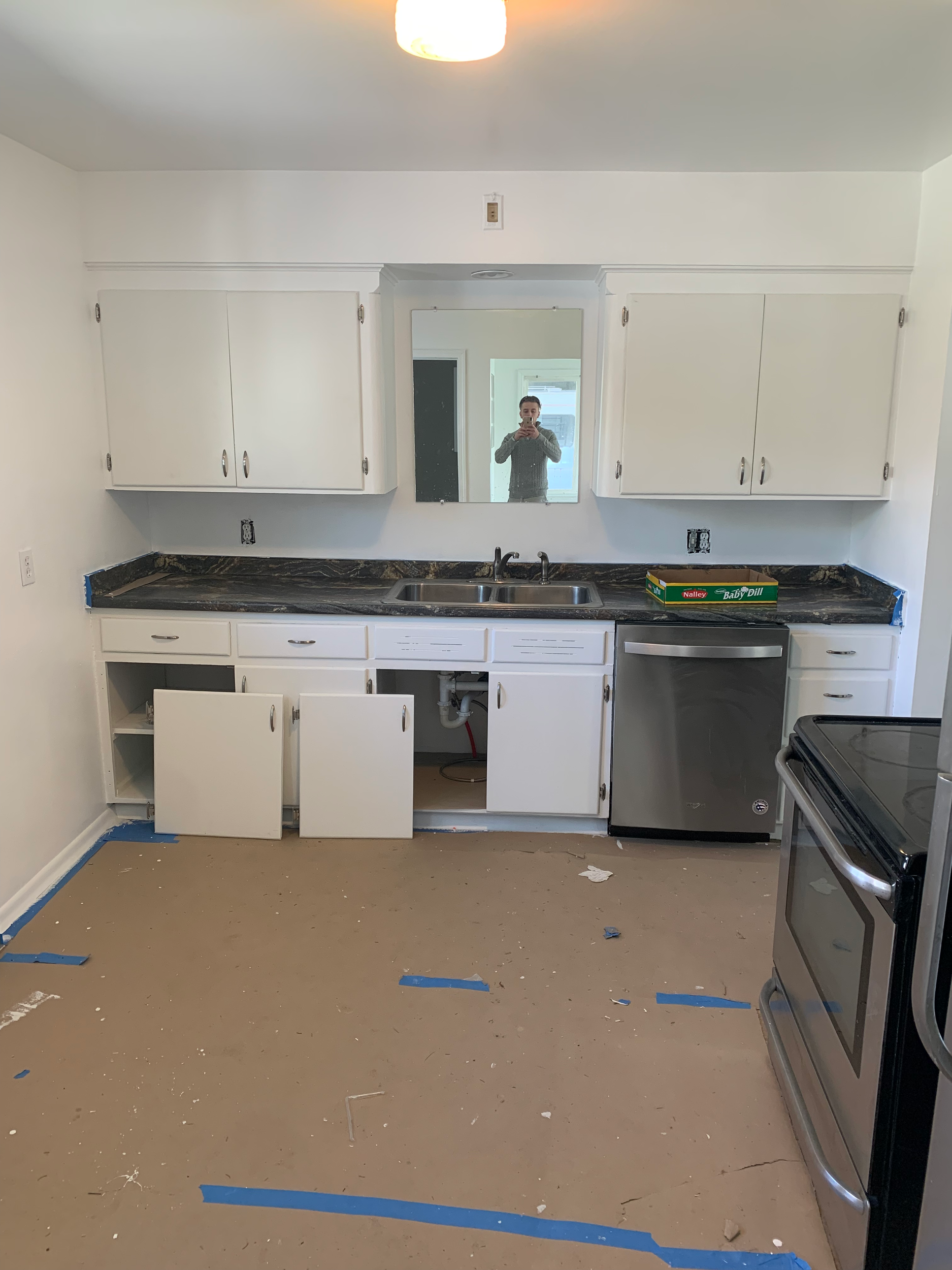 A partially renovated kitchen with white cabinets, a black countertop, dishwasher, and stove. The floor is covered with protective paper and blue tape. Some cabinet doors are open and there is an exposed sink area.