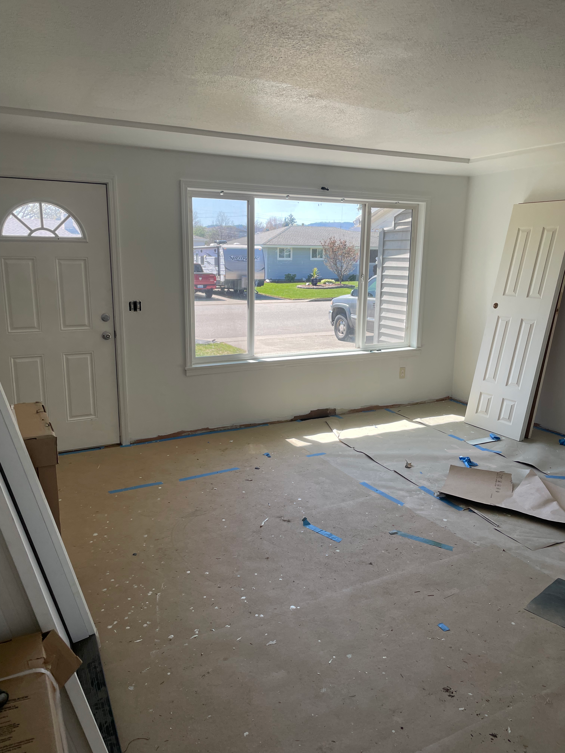 Interior room under renovation with bare floor, white walls, and covered windows. A door is propped against the wall. Outside view shows cars and houses.