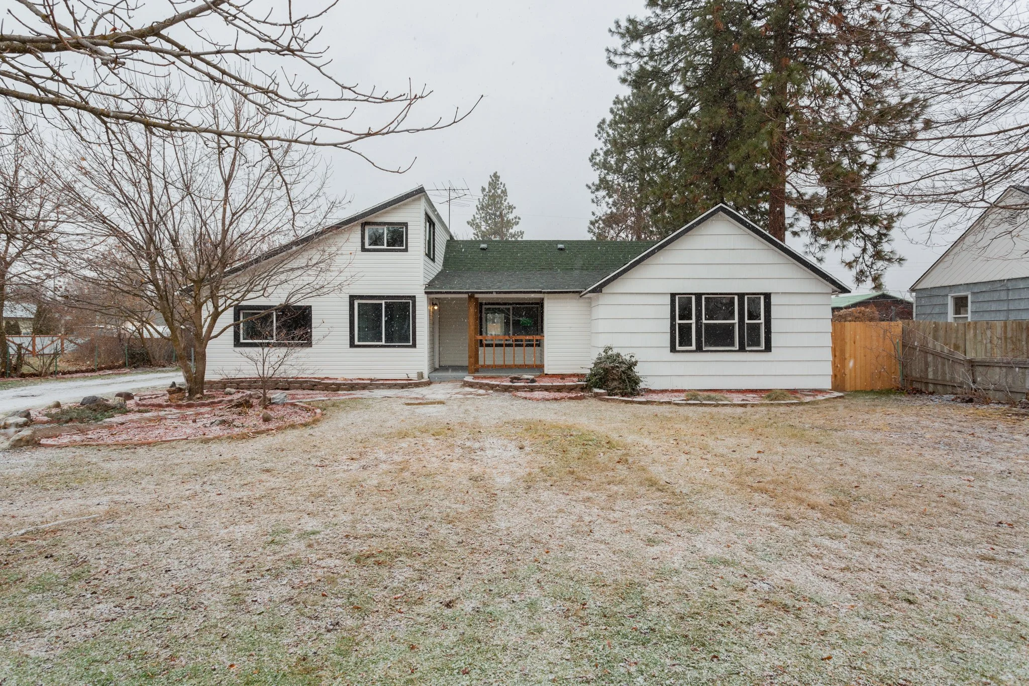 White suburban house with black trim in winter, surrounded by bare trees and a light snowfall, with a grassy yard and wooden fence.