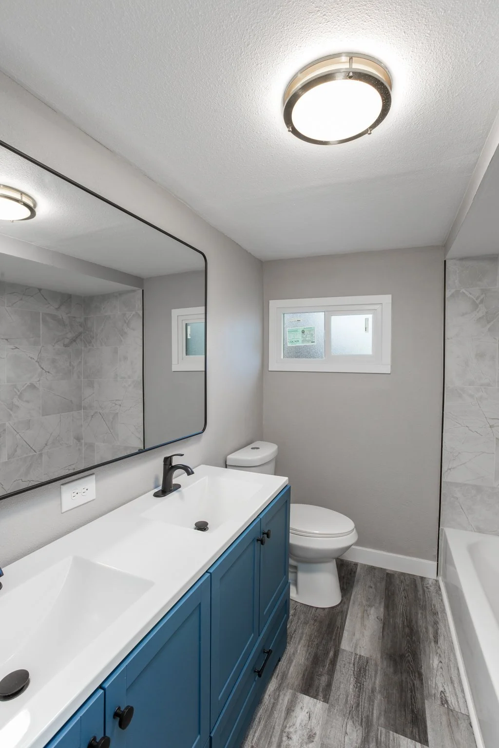 Modern bathroom with blue vanity, double sink, large mirror, toilet, and bathtub, featuring gray tile and wood-like flooring.
