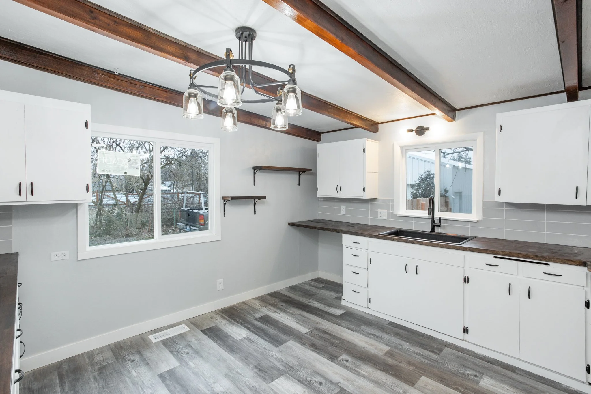 Modern kitchen with white cabinets, stainless steel sink, dark countertops, wooden ceiling beams, light gray tile backsplash, grey wood-look flooring, and a rustic chandelier.