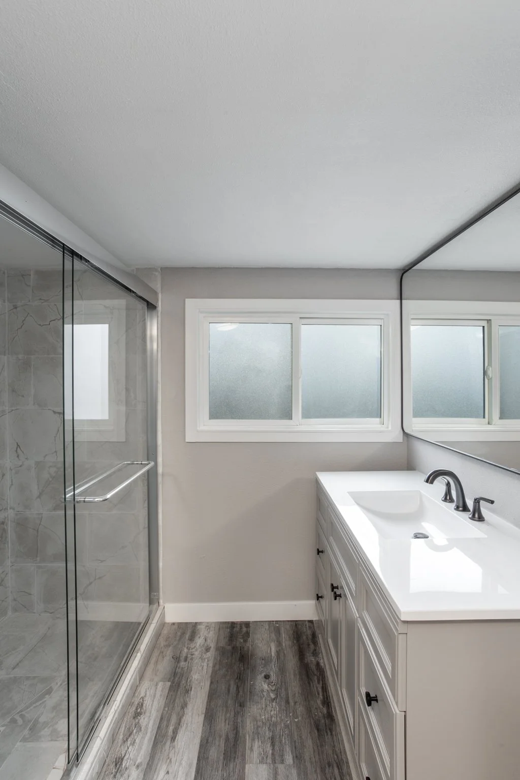 Modern bathroom with glass shower, vanity sink, mirror, and wood floor.