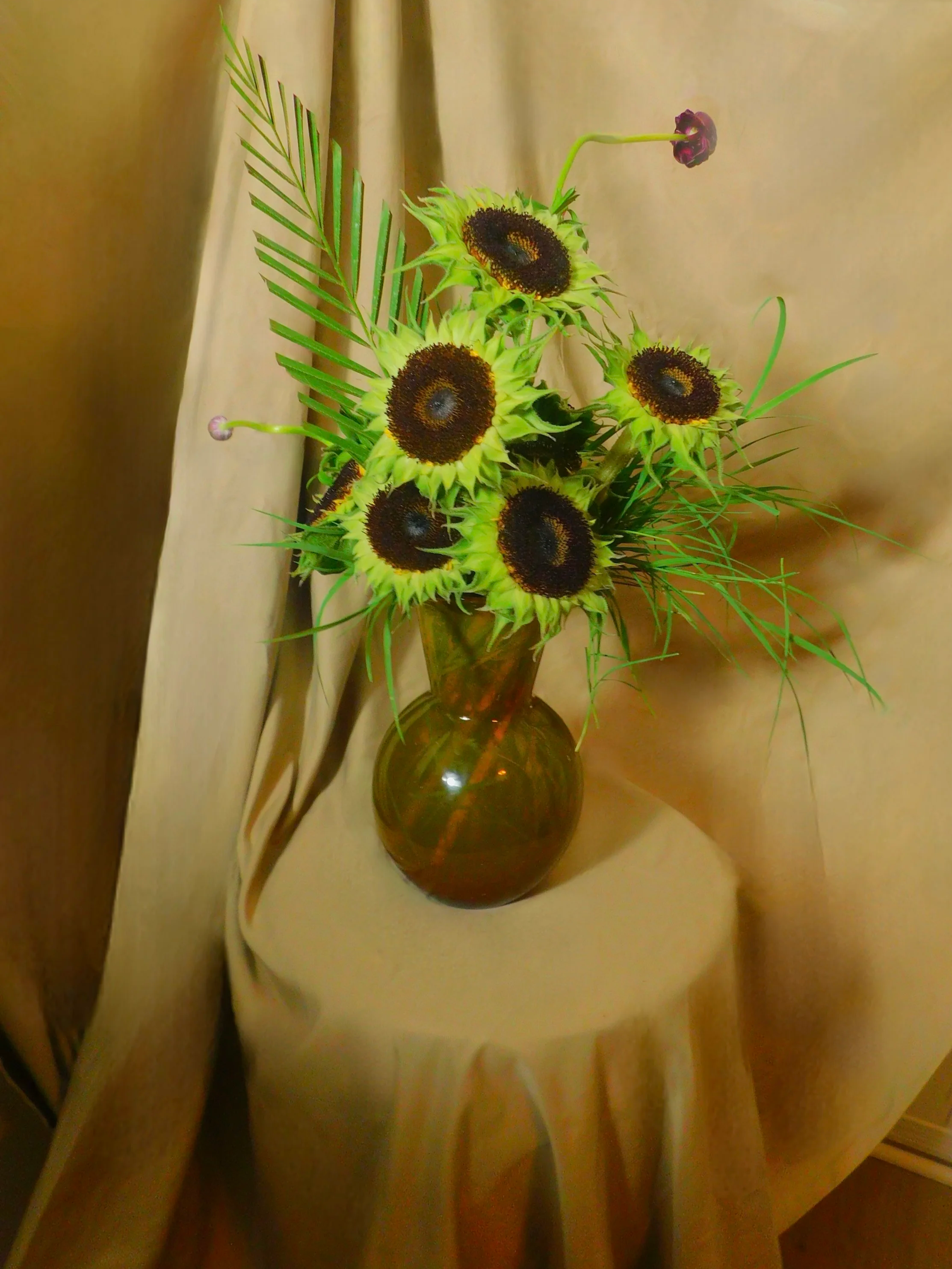 Vase containing sunflowers with dark centers, surrounded by green foliage and decorative leaves, set against a beige fabric backdrop.
