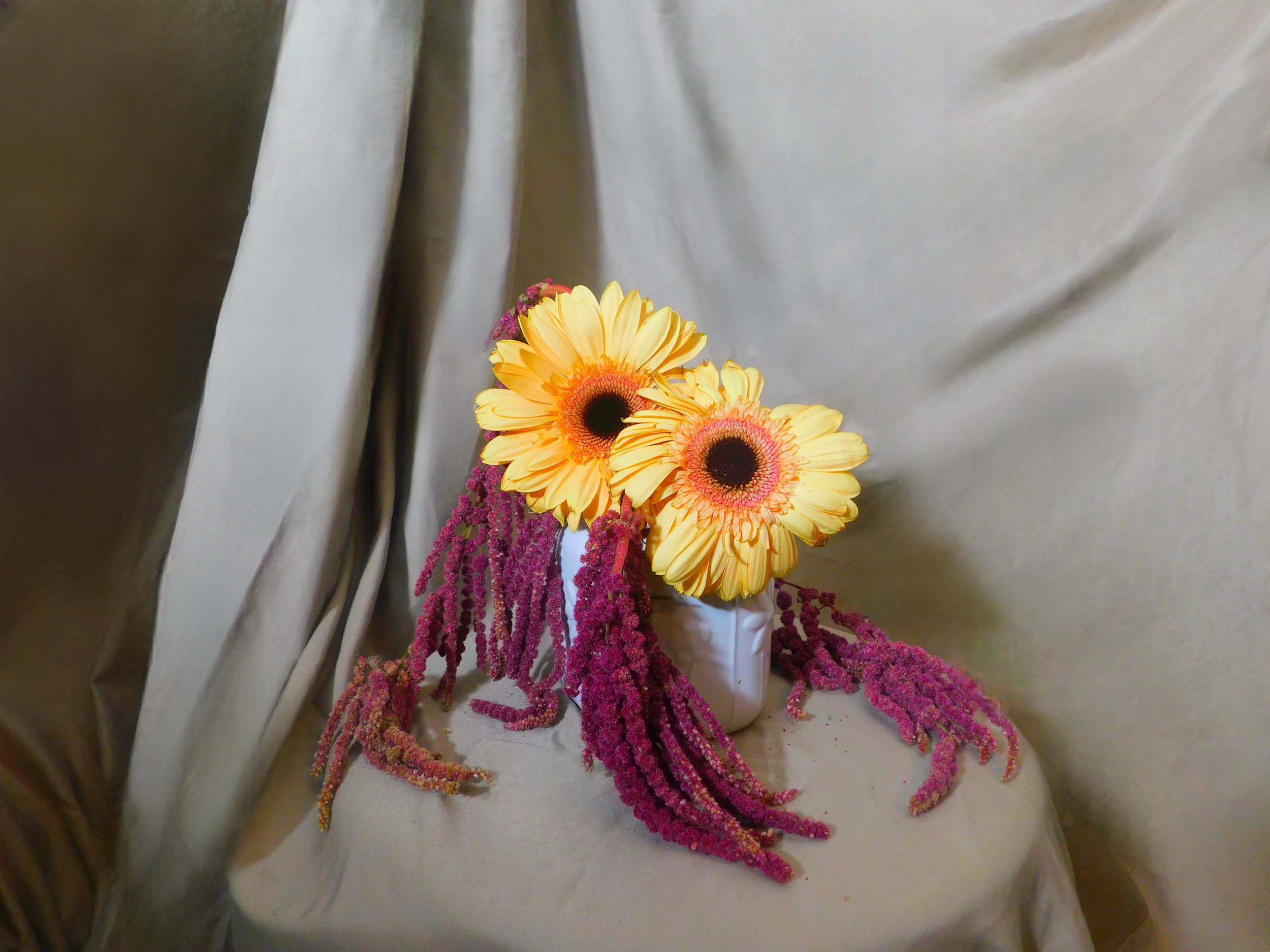 A bouquet of bright yellow gerbera daisies with dark centers, surrounded by pink and purple flowers, placed in a white vase on a neutral-colored surface with shiny gray curtains in the background.